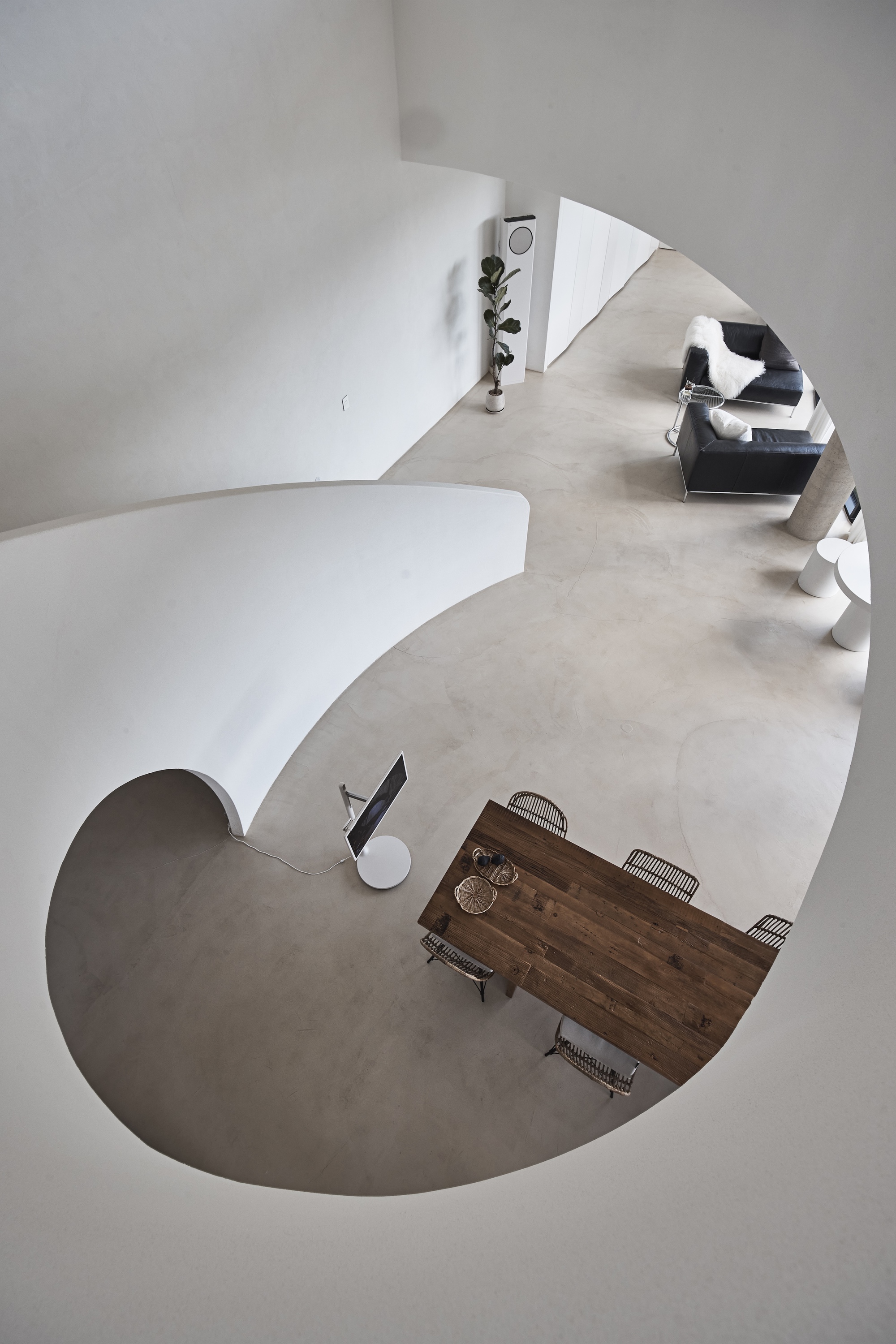 Overhead view through a curved white opening onto a timber dining table, wicker chairs and black sofas on a polished concrete floor