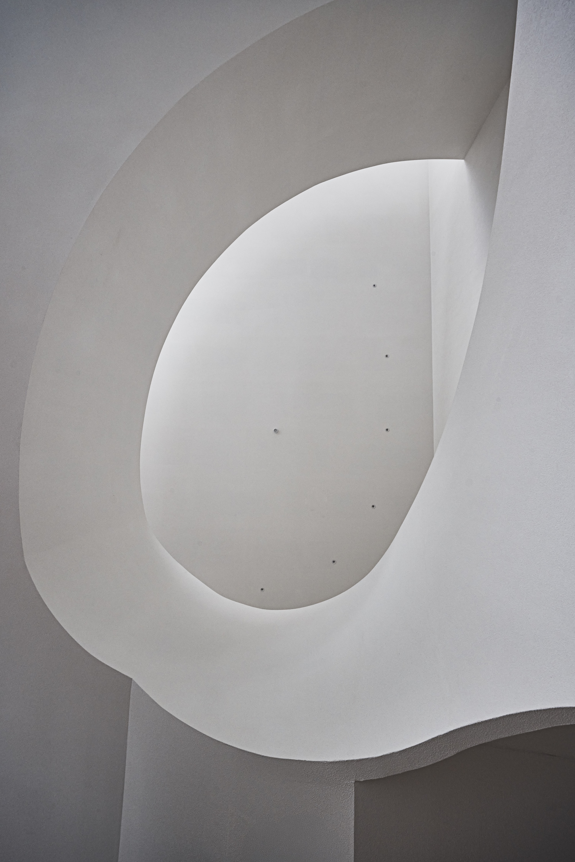 Looking up at a smooth white plaster oval opening with a looping curved edge, lit by soft daylight from above