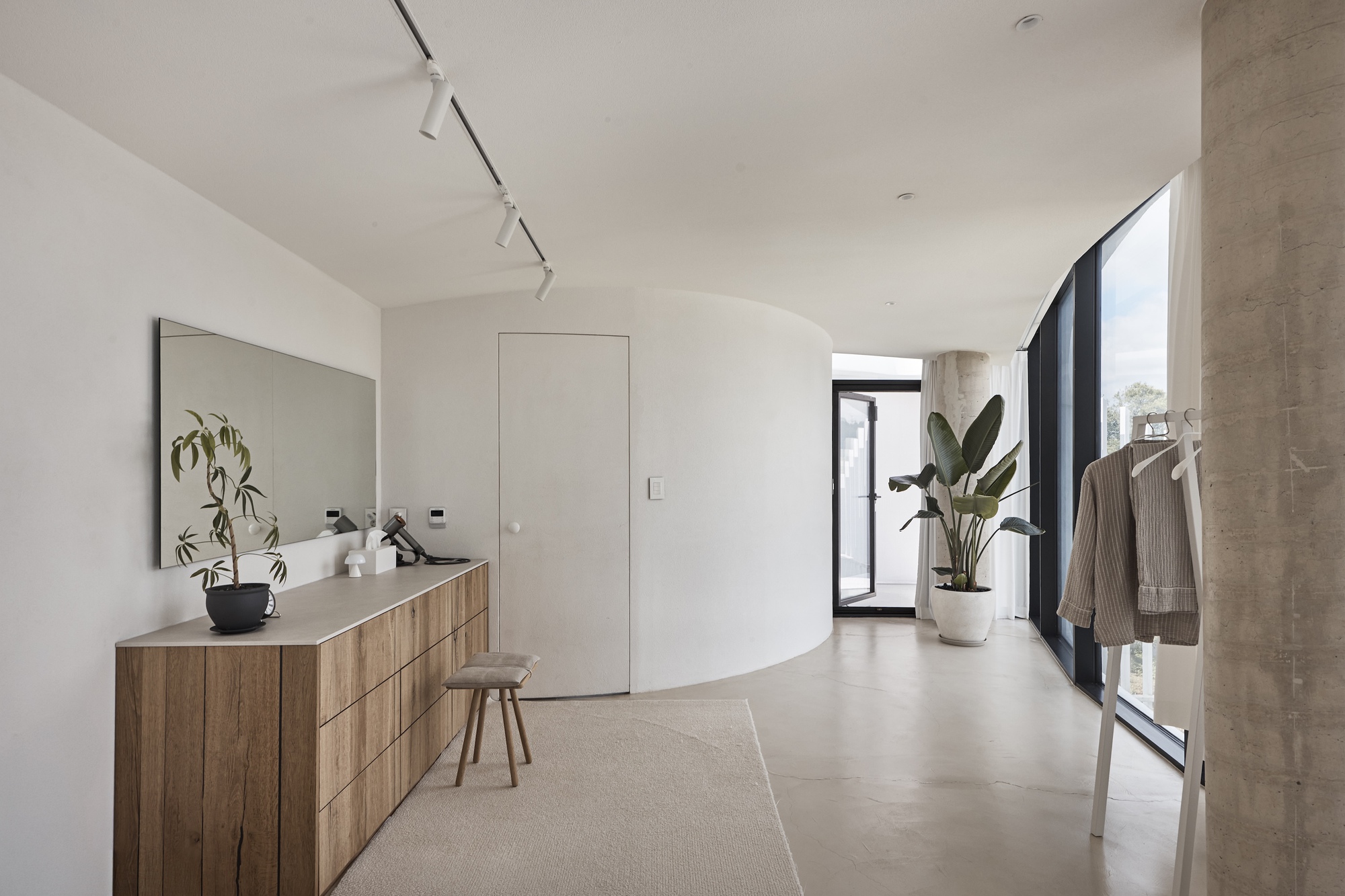 Entry hall with an oak drawer unit, long mirror and curved white wall, beside a concrete column and tall window