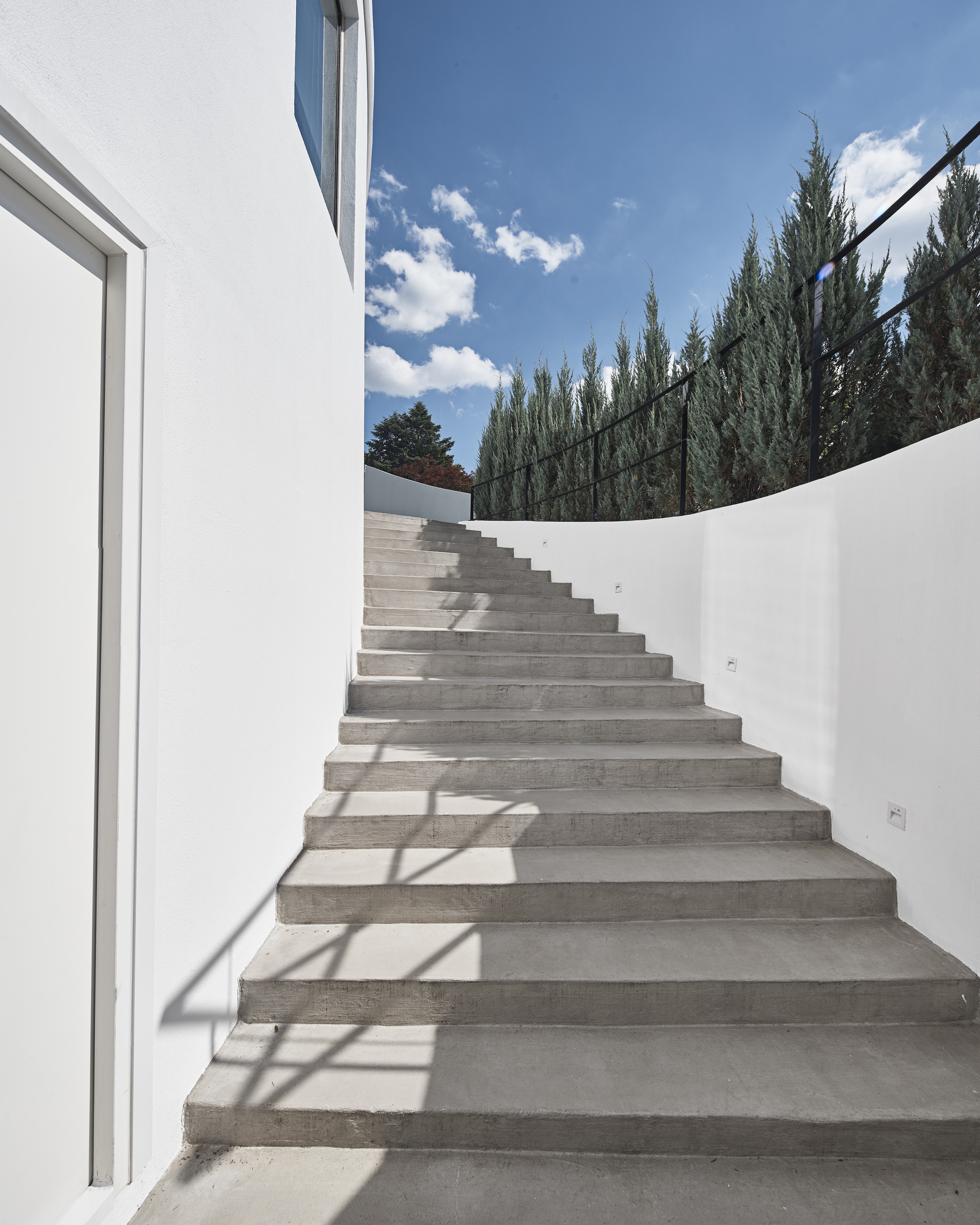 Concrete exterior stair rising between a curved white wall and a black railing, with cypress trees against a blue sky