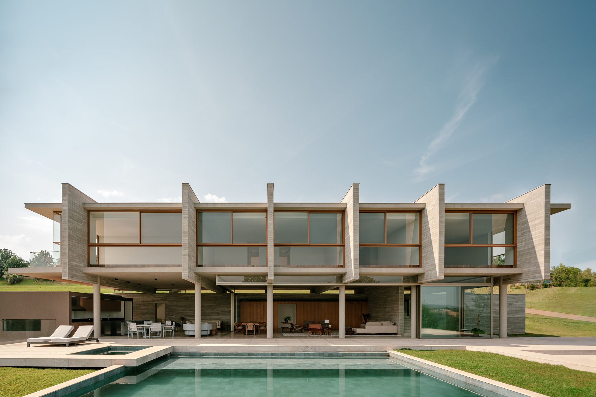 Front elevation of the two-storey house with angled board-formed concrete fins and timber-framed glazing above a pool, under a clear blue sky