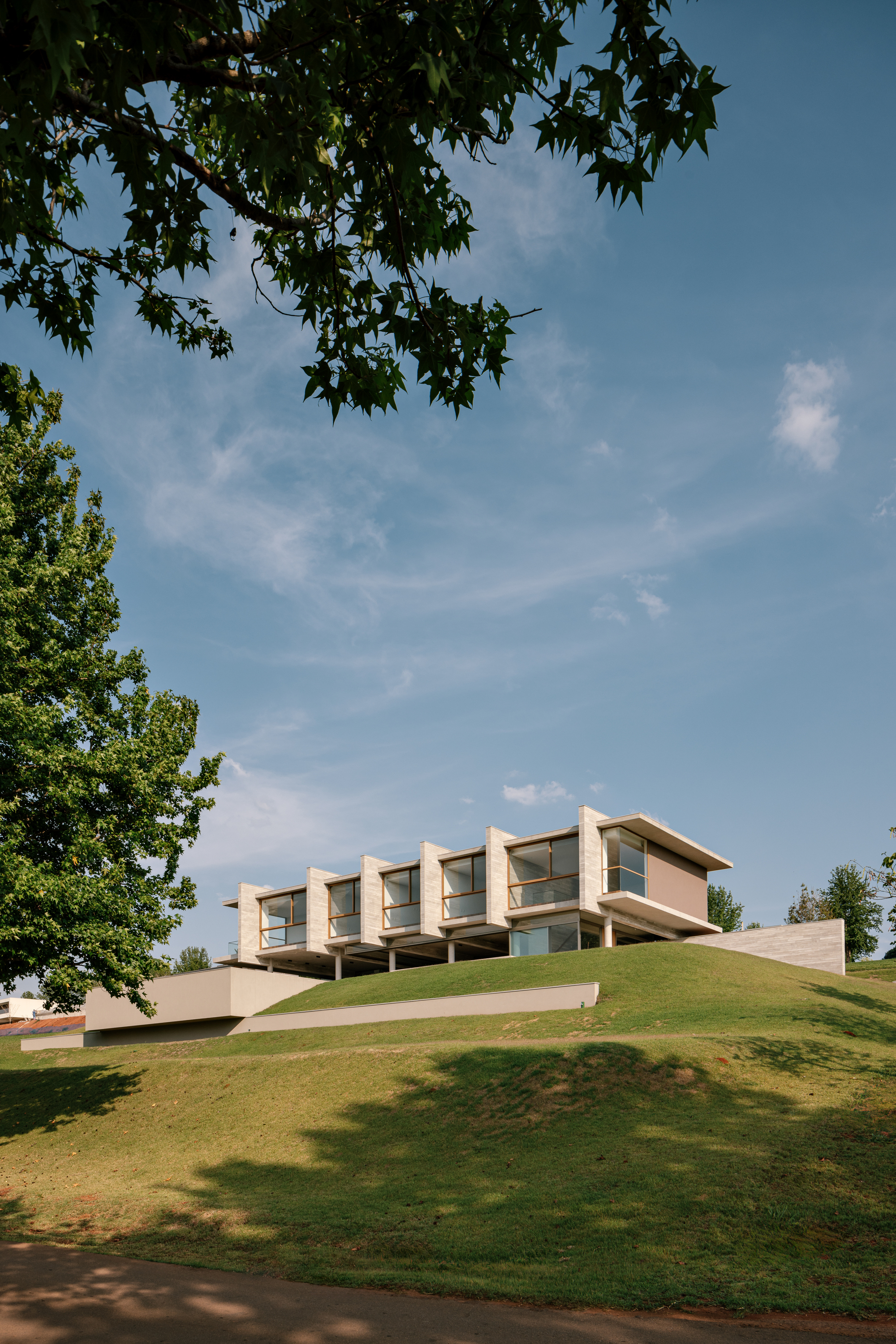 House raised on a grassy mound, seen from below through the dark leaves of a tree in full sun