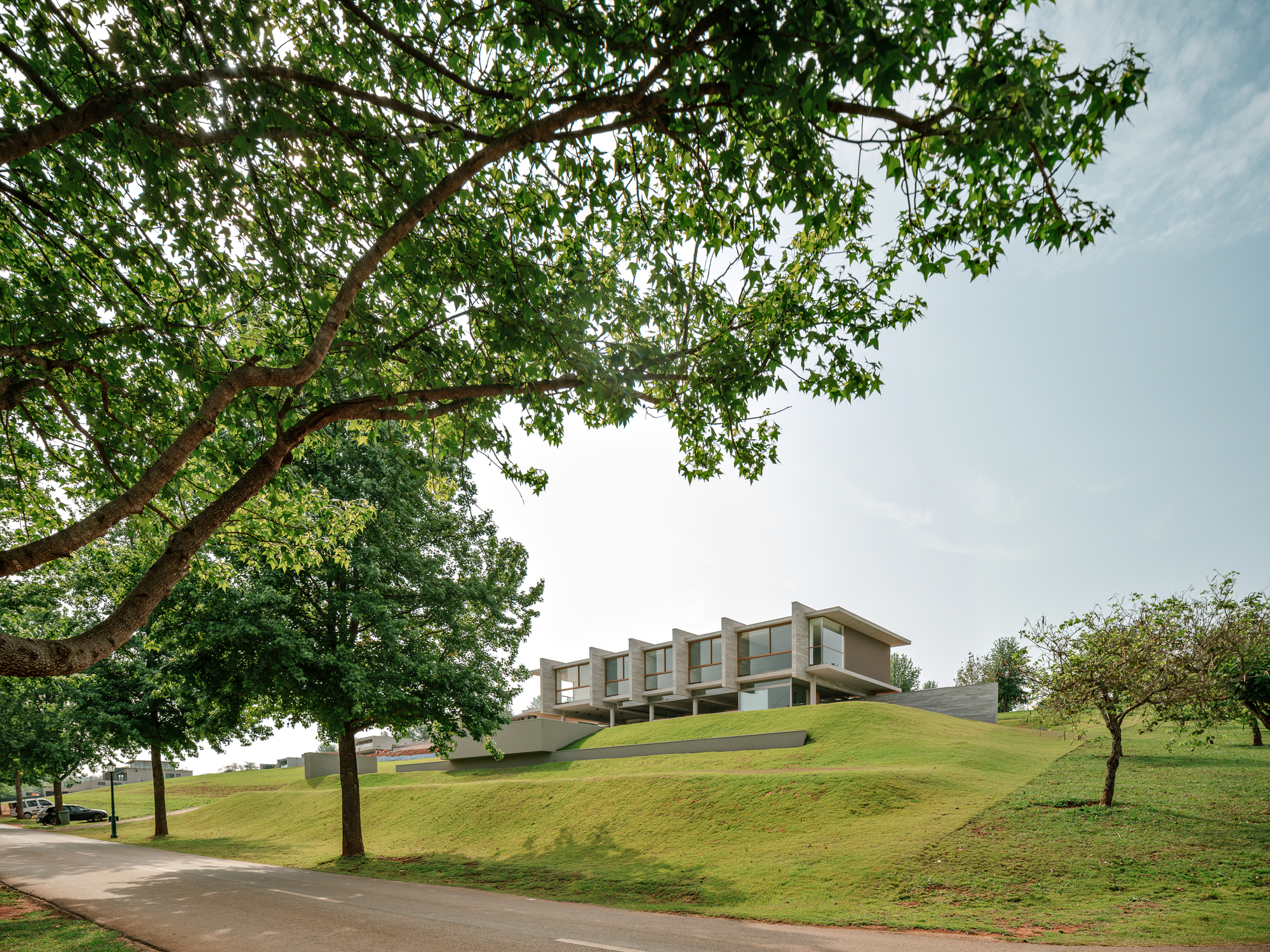 House on its landscaped hill seen across a curving road, framed by a large tree and small orchard trees