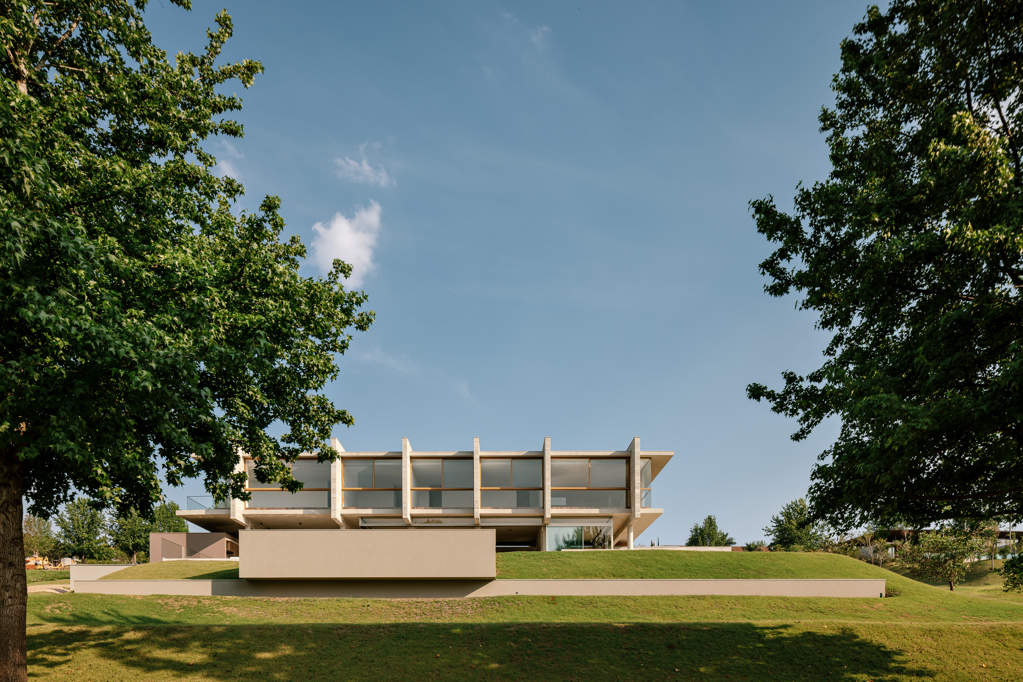 Two-storey house with slender vertical fins and a long glazed band, set above a grassy slope between two leafy trees