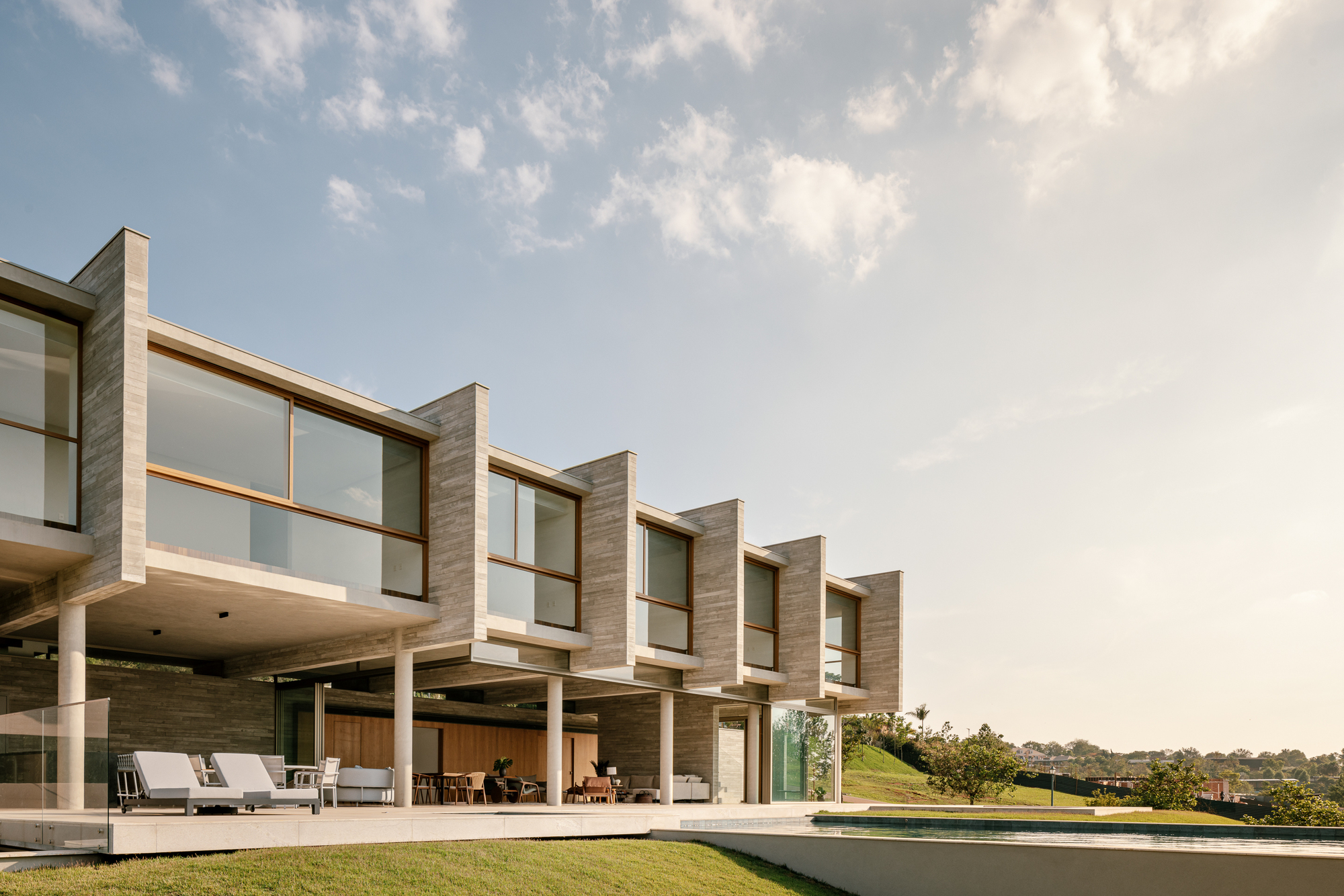 Stepped board-formed concrete bays with timber-framed windows over a covered terrace, lit by low evening sun beside the pool