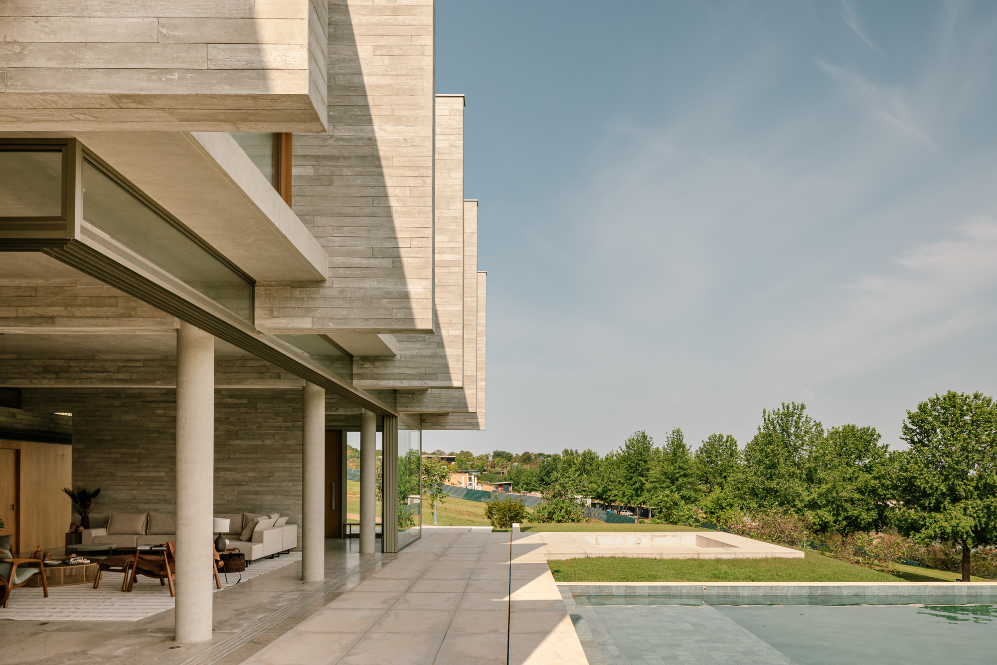 Covered terrace with round columns and textured concrete walls opening onto a pool edge and green hillside under clear sky