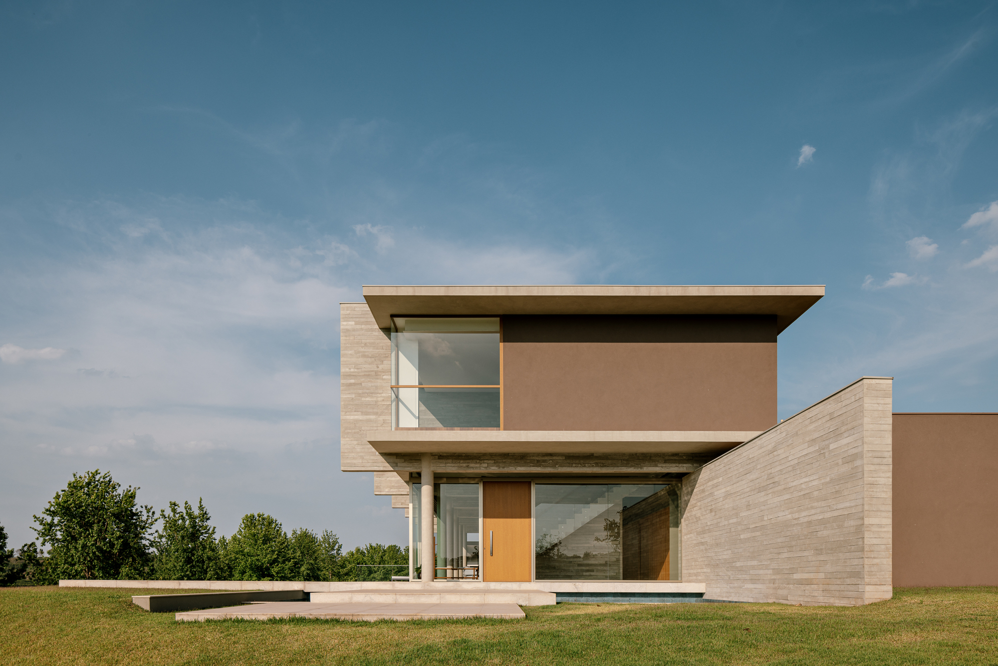Entrance facade with a timber door, cantilevered concrete slabs and a brown rendered wall beside a board-marked stone wing