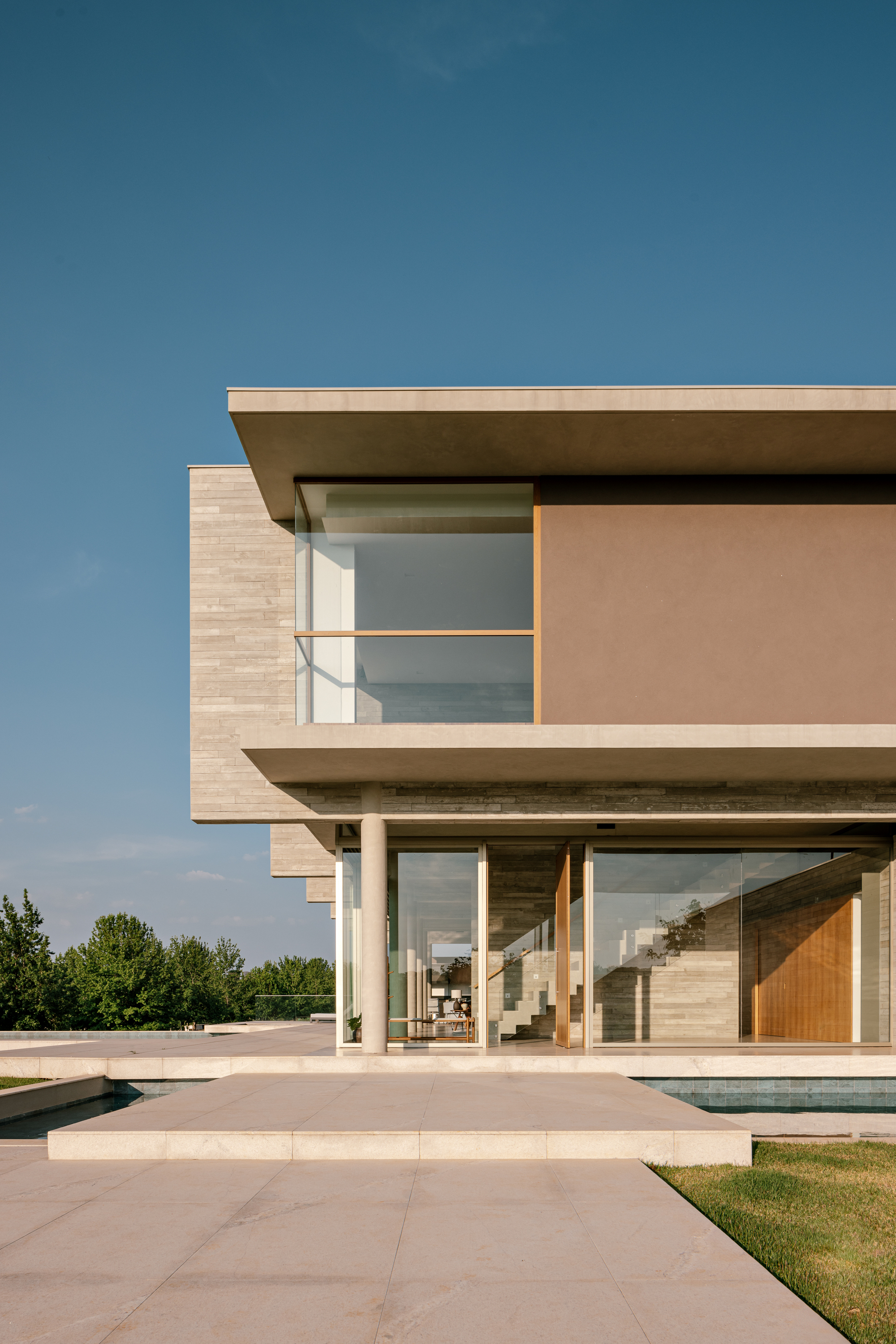 Vertical view of the glazed ground floor under projecting slabs, with a stair visible inside and a shallow pool in front