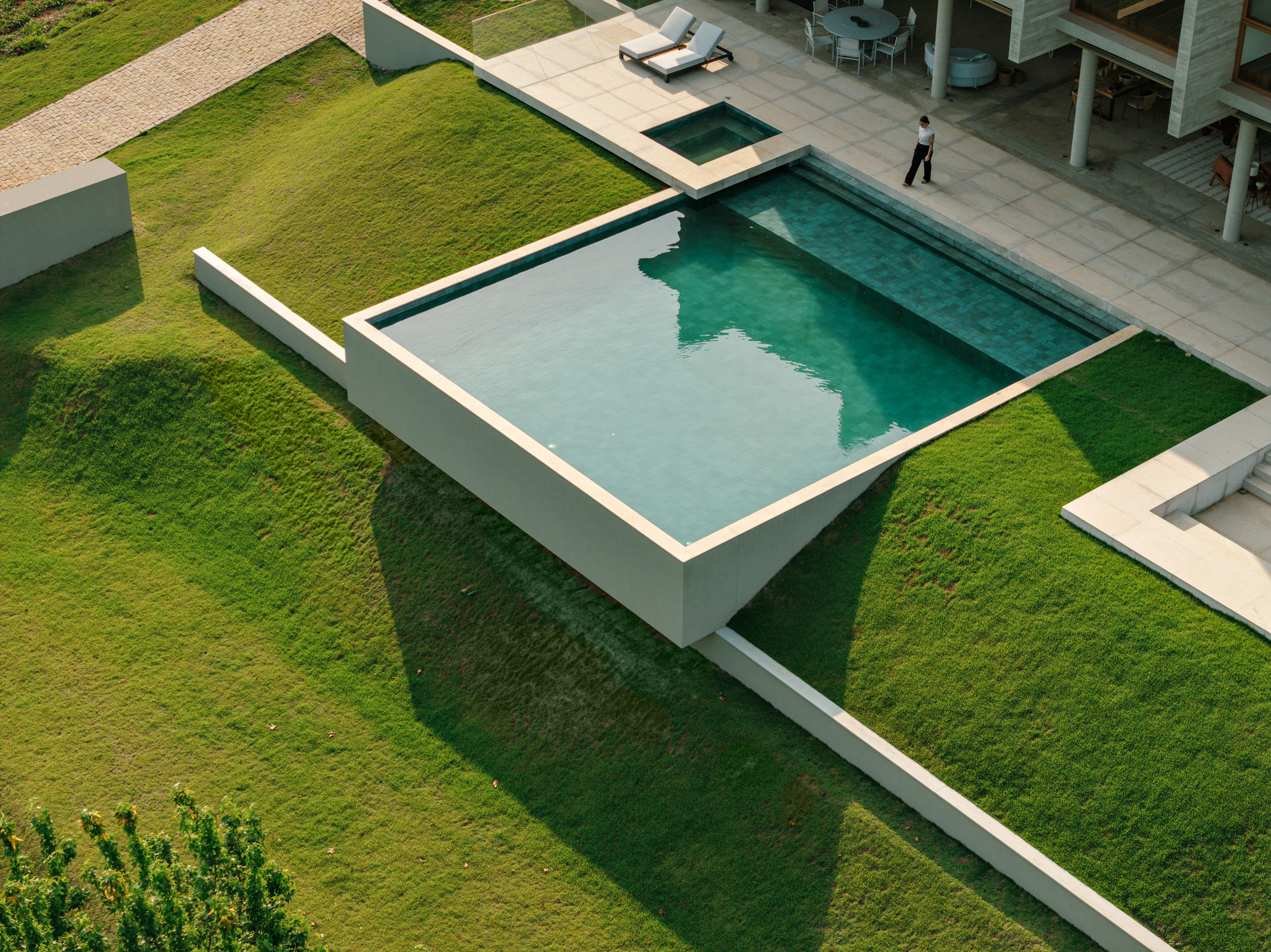 Aerial view of a square pool cantilevered over a grassed slope, with a person walking across the paved terrace at dusk