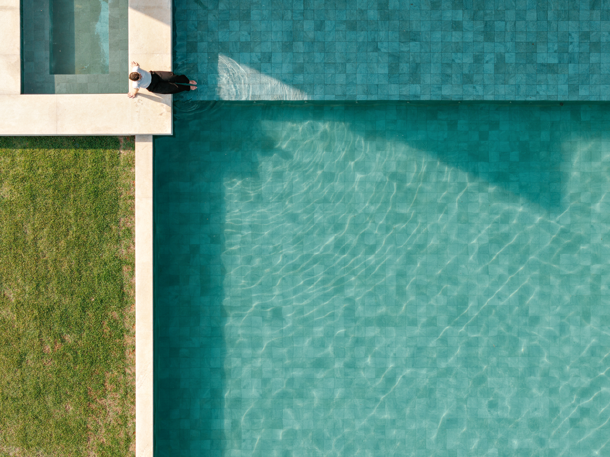 Top-down view of a green-tiled pool edge with a person sitting at the corner beside a lawn in afternoon sun