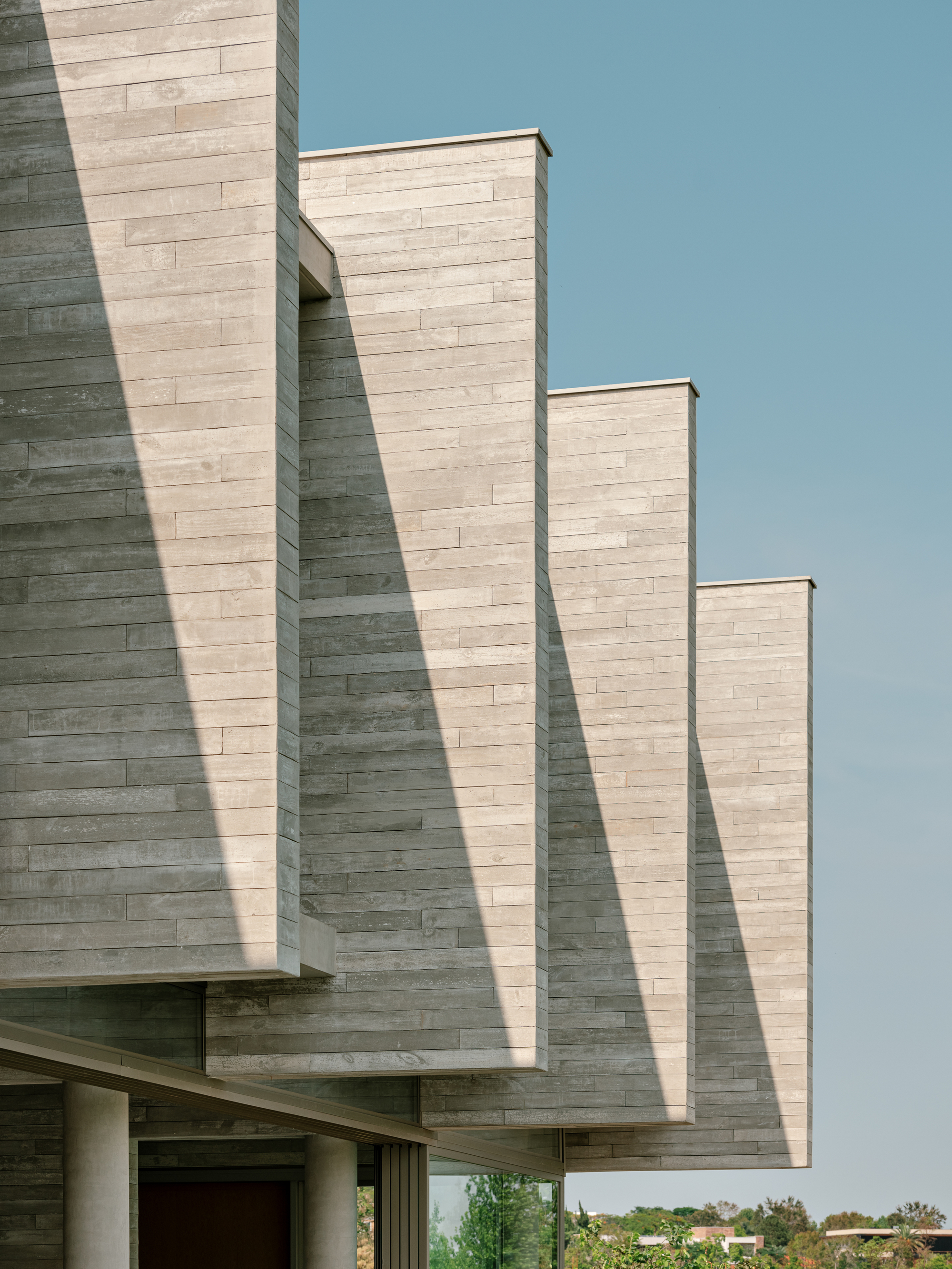 Stepped stone-clad volumes cantilevering over a glazed ground floor, casting long diagonal shadows under a clear blue sky