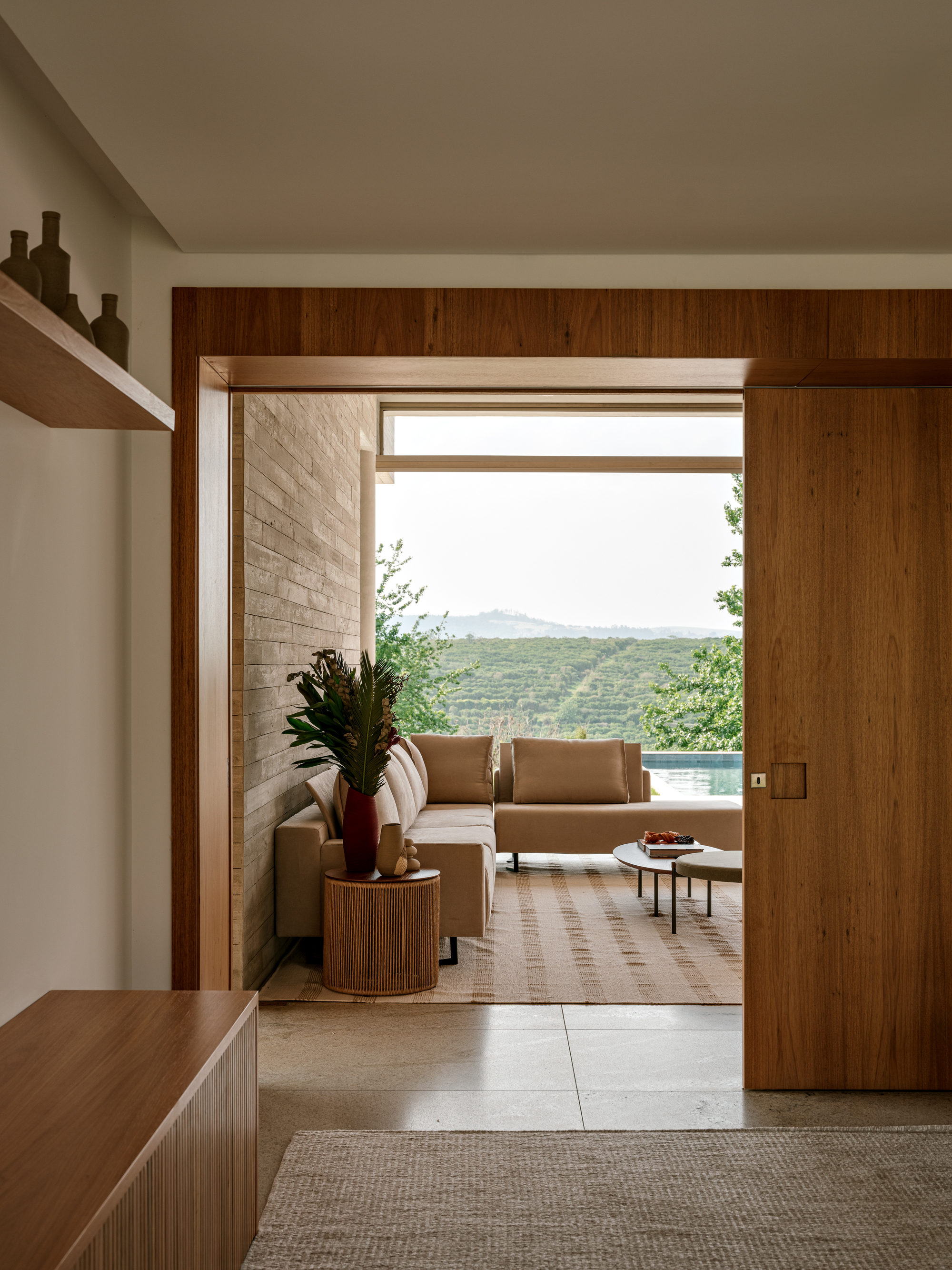 View through a wood-framed opening to a sofa on a covered terrace, with pool and planted hills beyond in hazy light