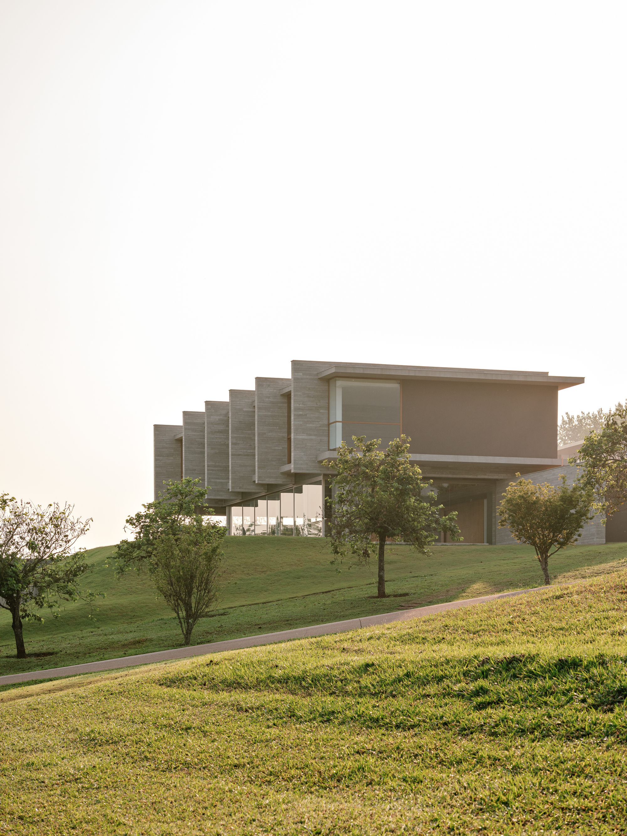 Board-formed concrete house with stepped cantilevered fins on a grassy rise, a curving path and young trees in morning haze