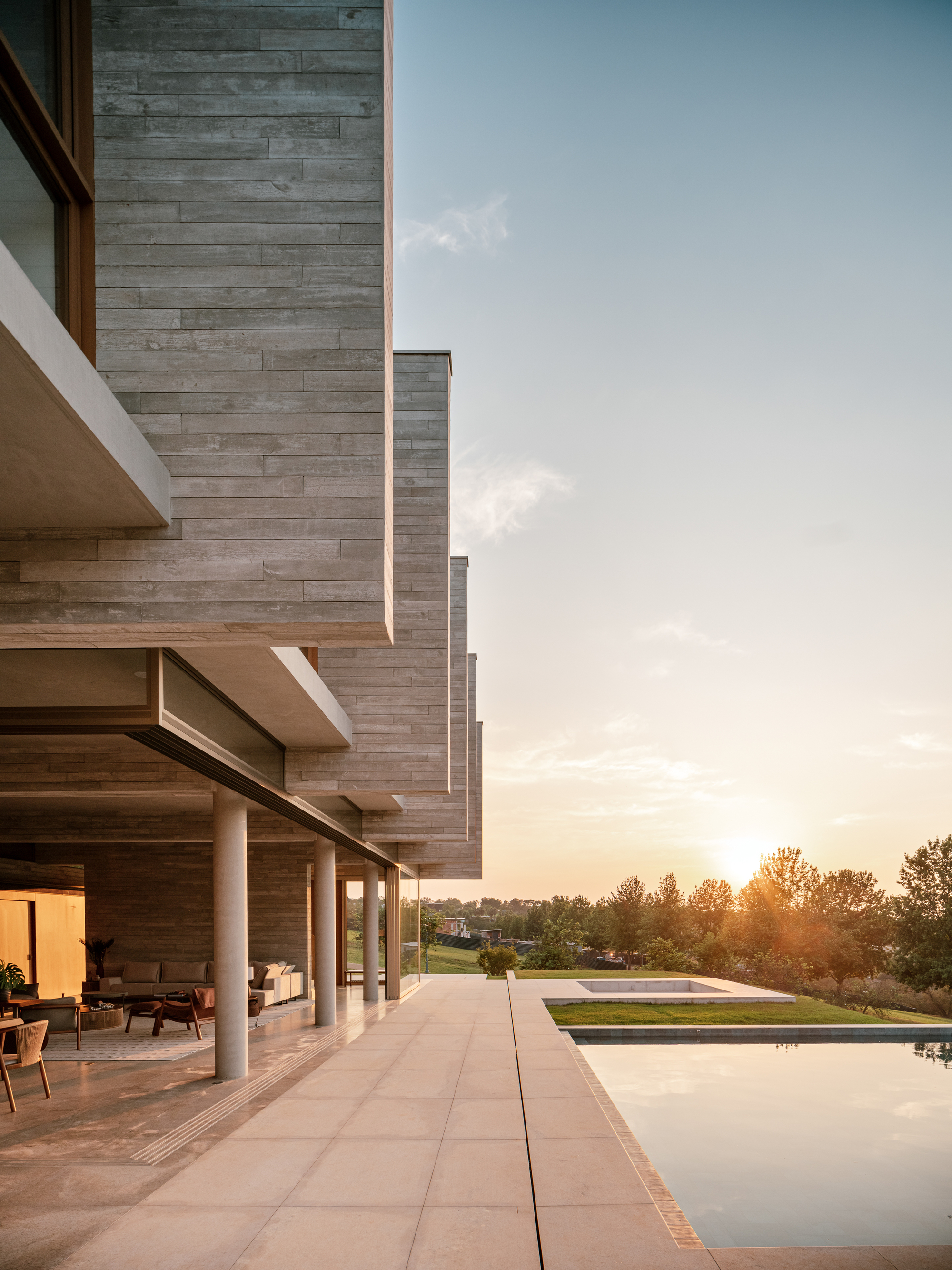 Covered terrace under stacked concrete fins with slim round columns, facing a pool and low sun at dusk