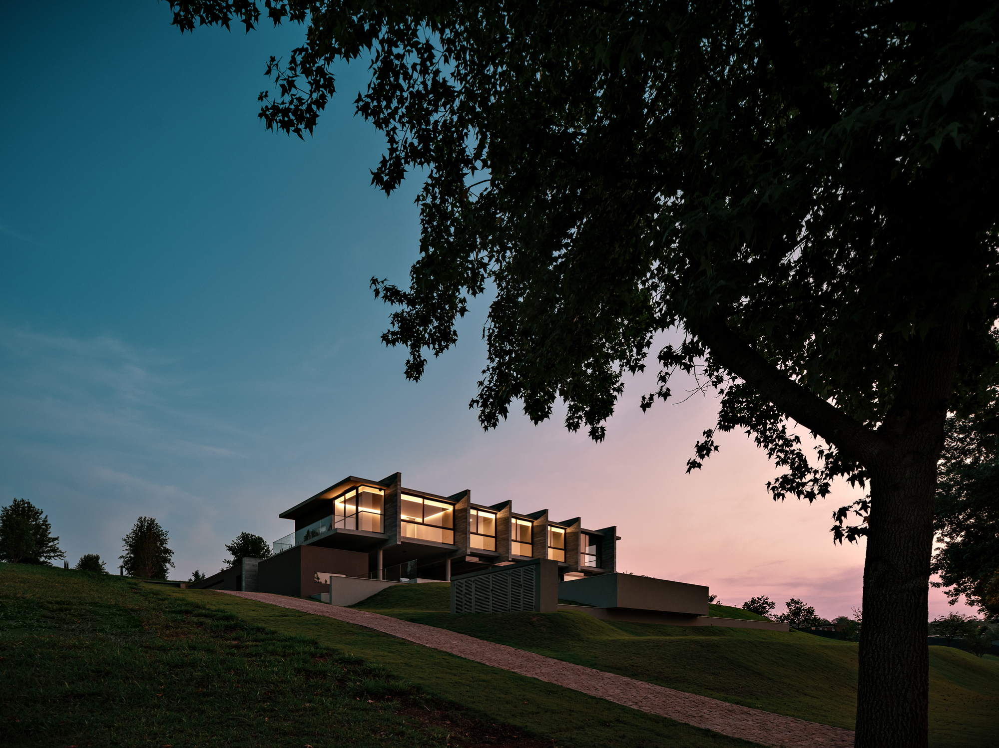Lit house with a row of angled concrete fins on a hilltop, framed by a large tree against a pink dusk sky