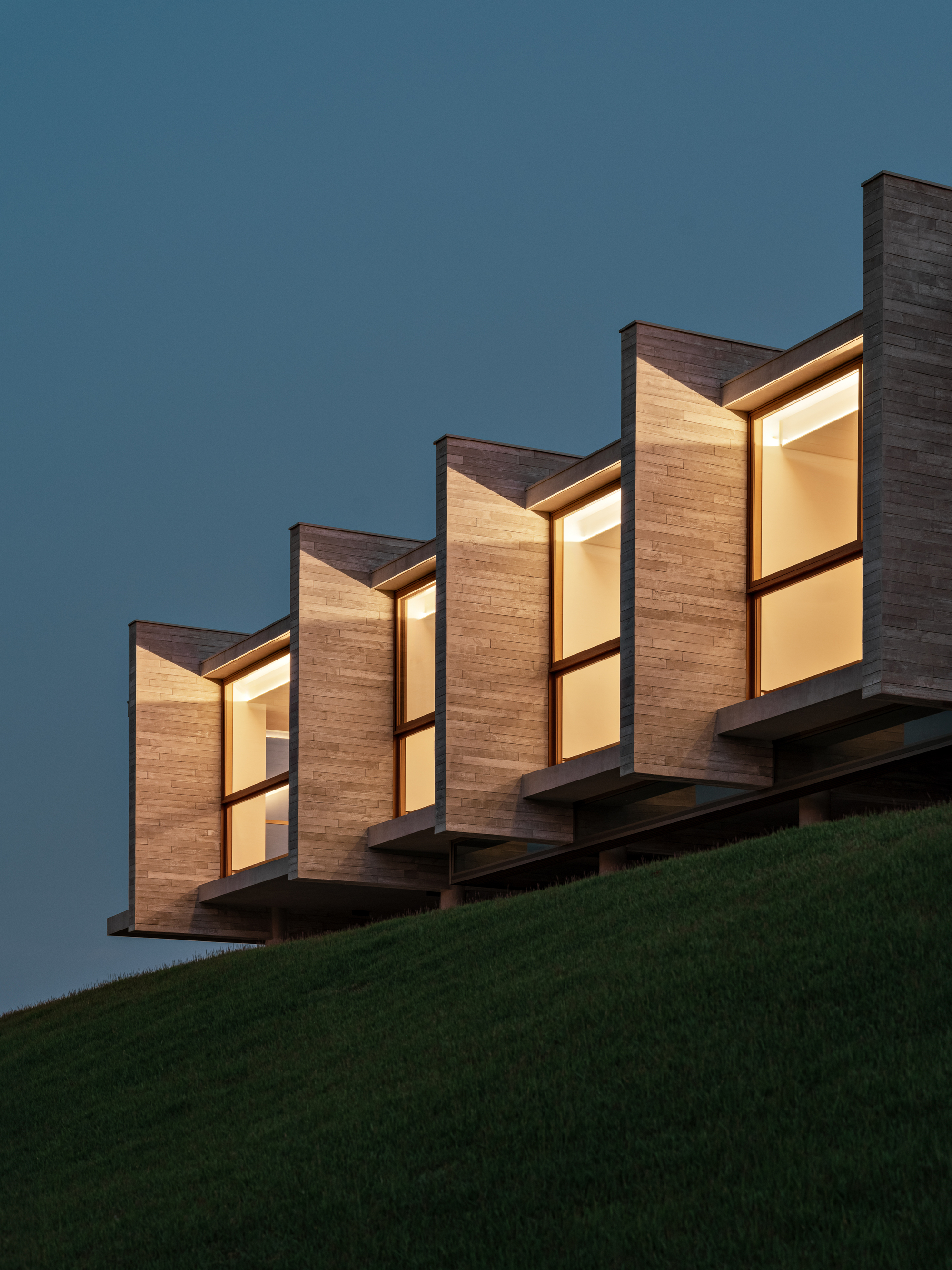 Close view of the board-formed concrete fins and glowing timber-framed windows cantilevered over a dark grass slope at twilight