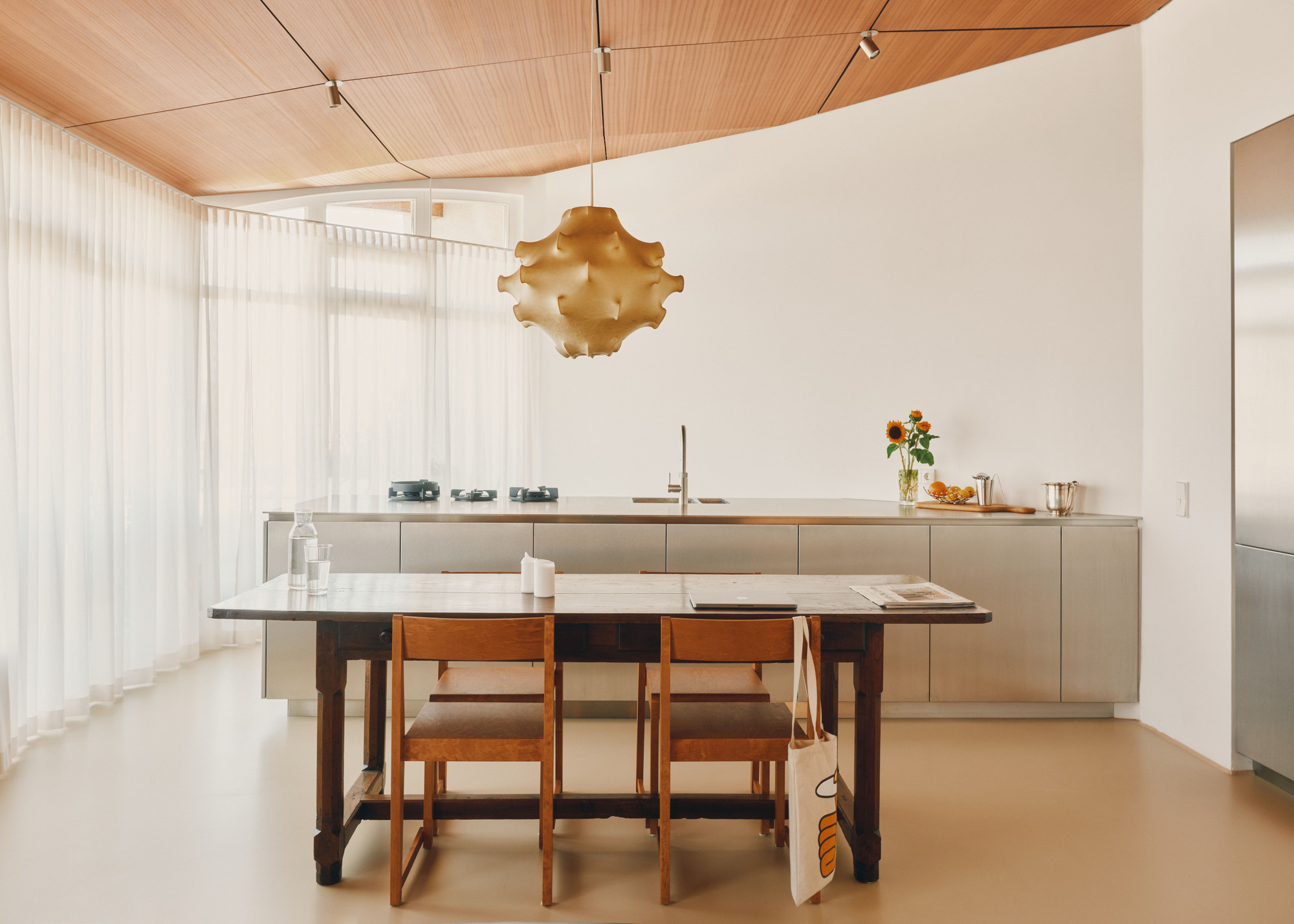 Long timber dining table and wooden chairs in front of a stainless steel kitchen counter, with sheer curtains filtering daylight