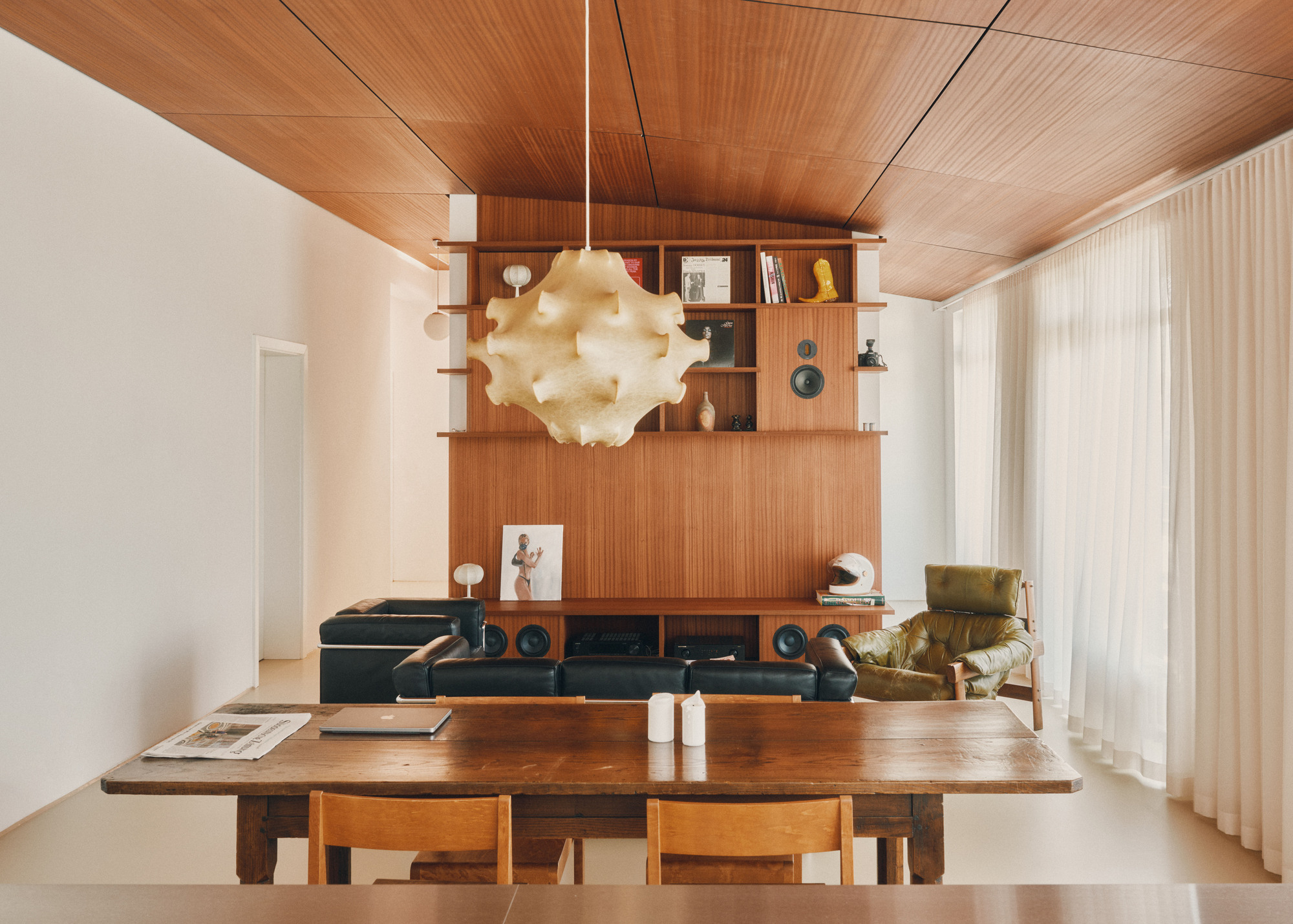 Teak wall unit with shelves and speakers behind a black leather sofa, under a slanted veneer ceiling and a paper pendant lamp