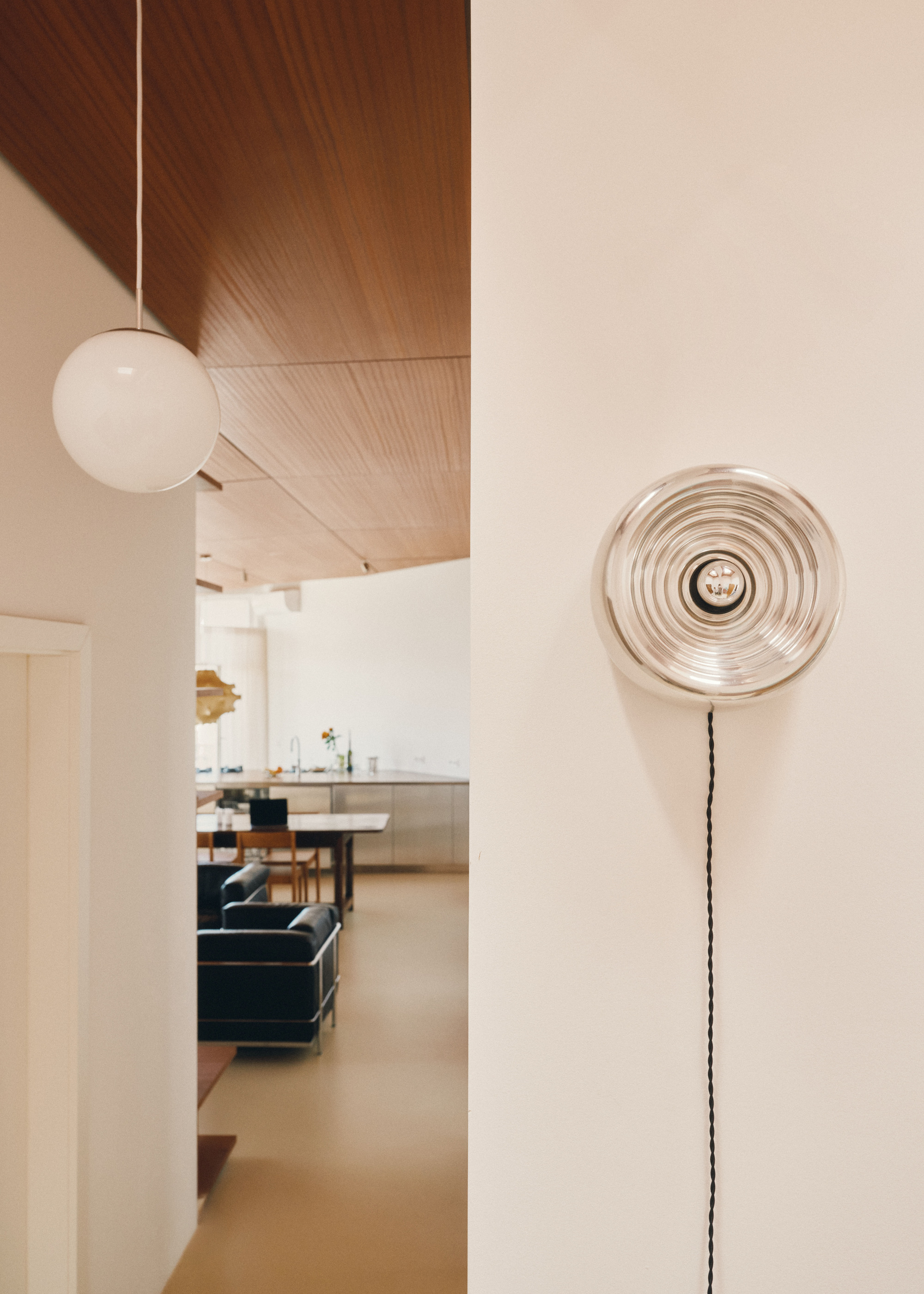 Doorway view past a round glass pendant and ribbed wall light toward a timber ceiling, leather armchairs and a steel kitchen