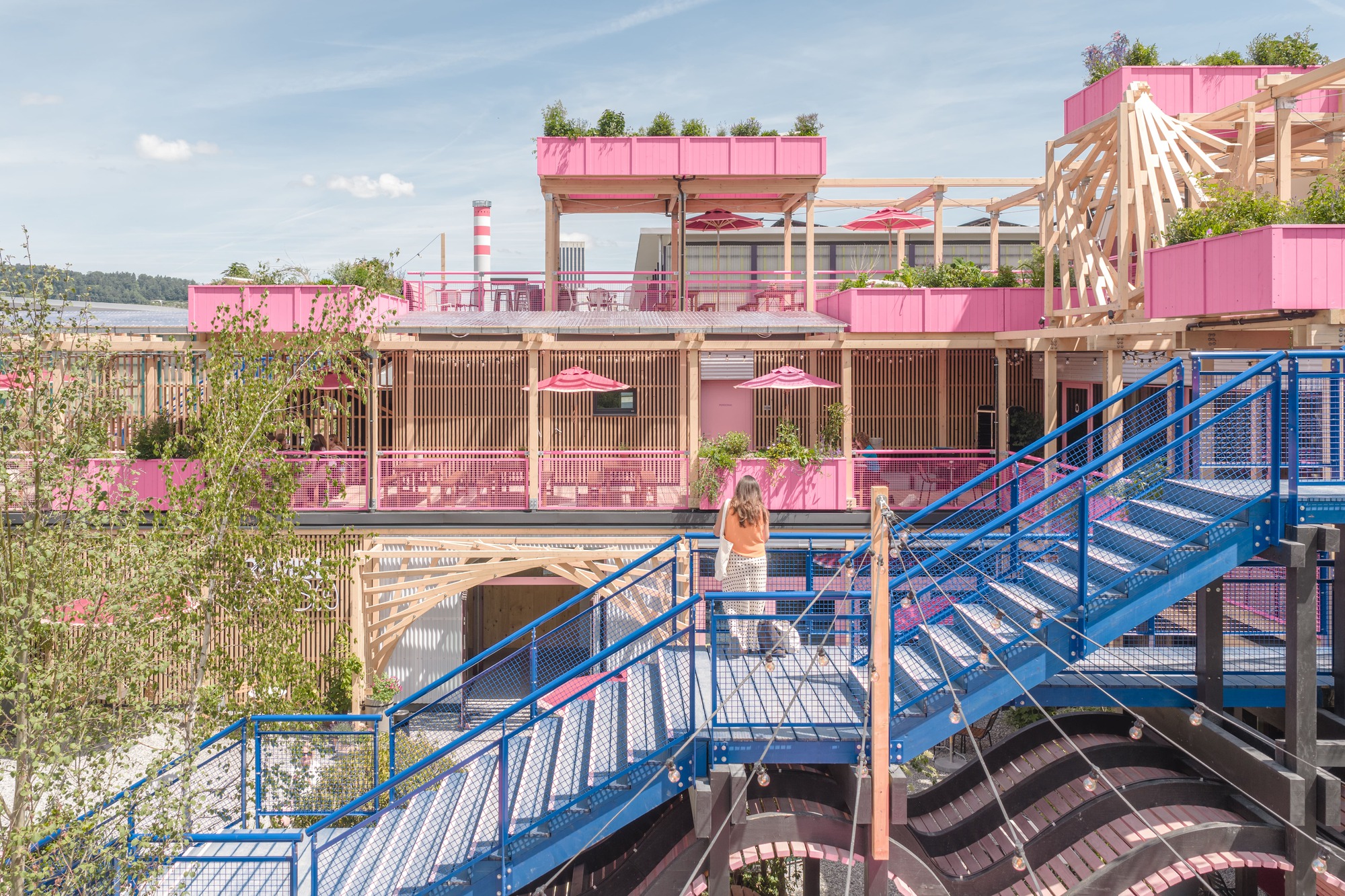 Blue steel stair with mesh balustrade climbing toward pink-painted timber terraces, with a woman standing on the landing under a bright summer sky