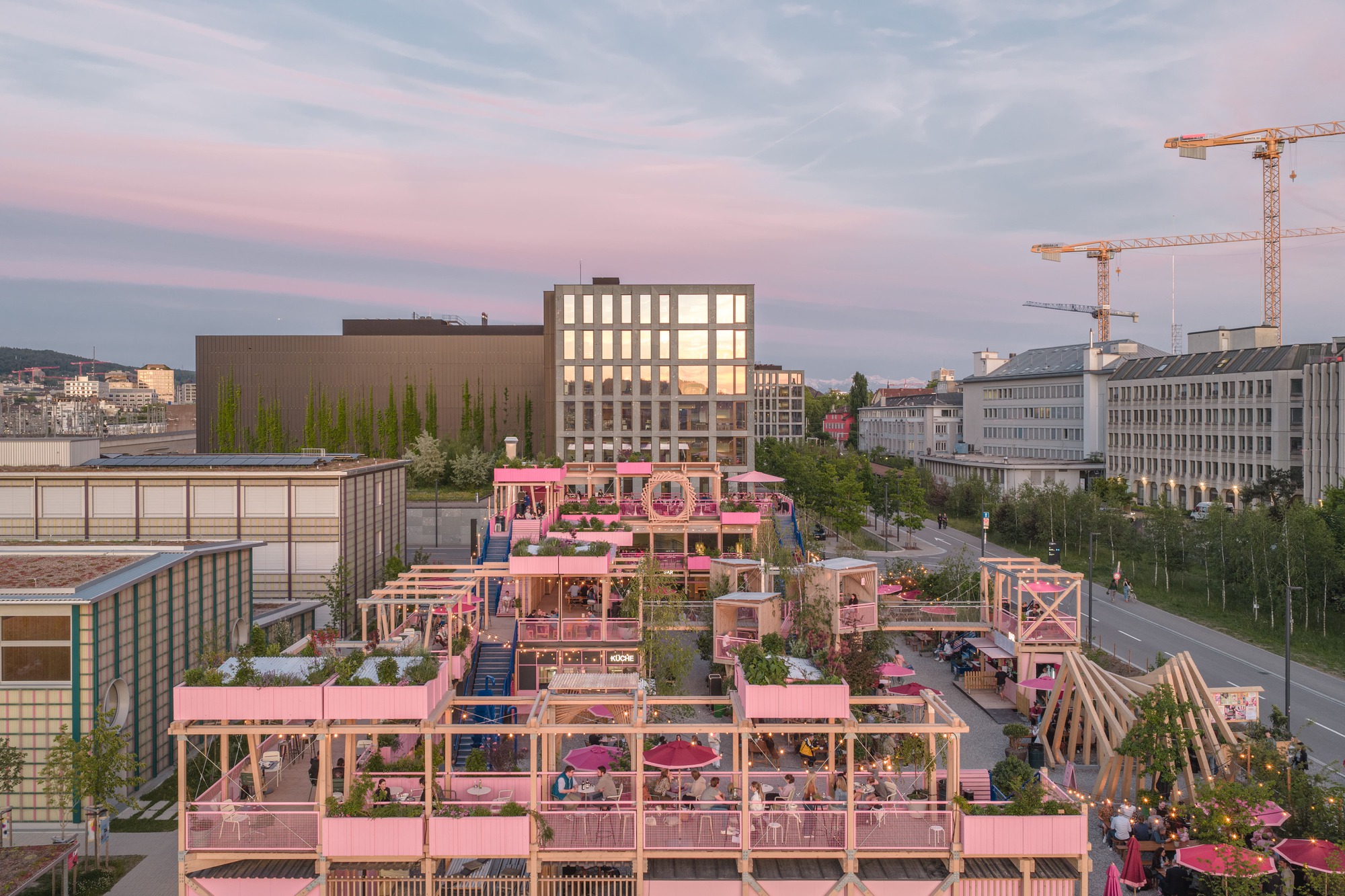Aerial view of stacked timber terraces with pink planters and umbrellas, framed by construction cranes at dusk under a pink sky