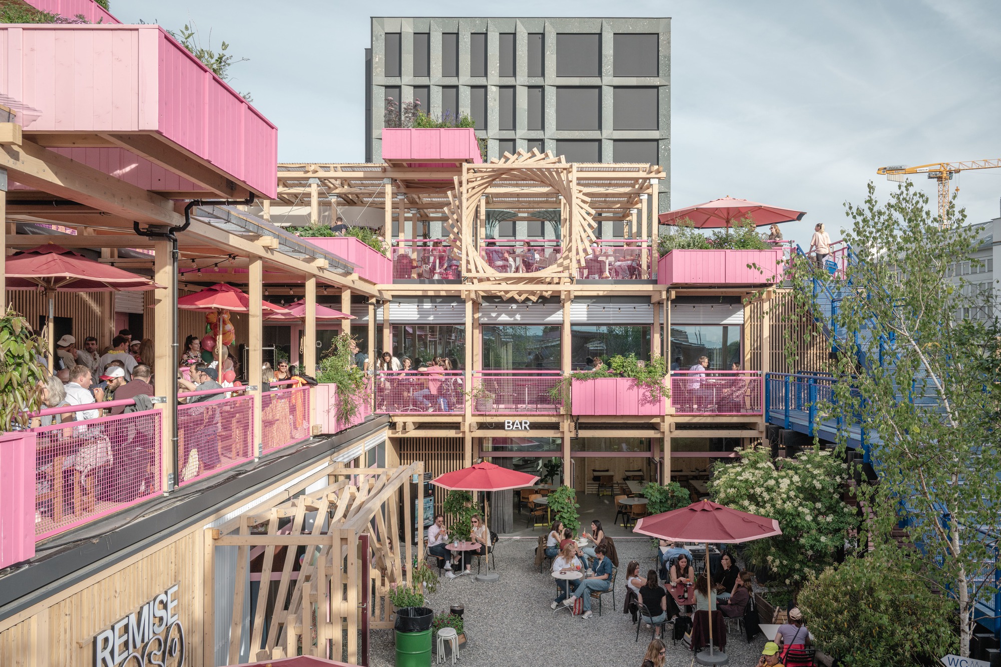 Courtyard bar facade of timber balconies and pink planters, with diners at tables below under red umbrellas and a flowering tree