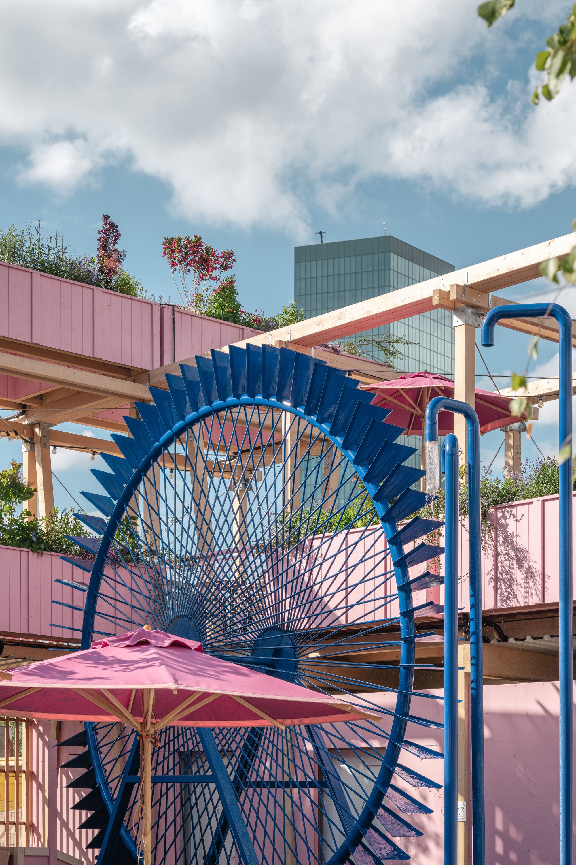 Blue steel wheel with radiating spokes beside a pink umbrella and pink timber walls, set against a cloudy sky