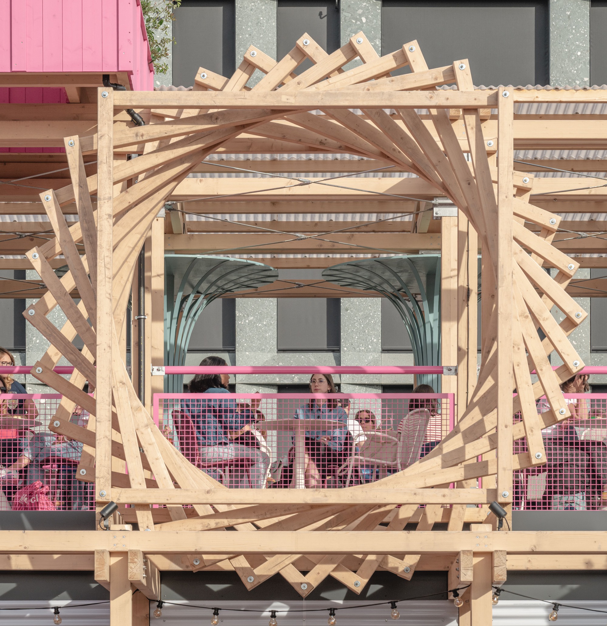 Square timber frame holding a spiral of crossed wooden slats, with diners seated behind pink mesh railings in warm evening light