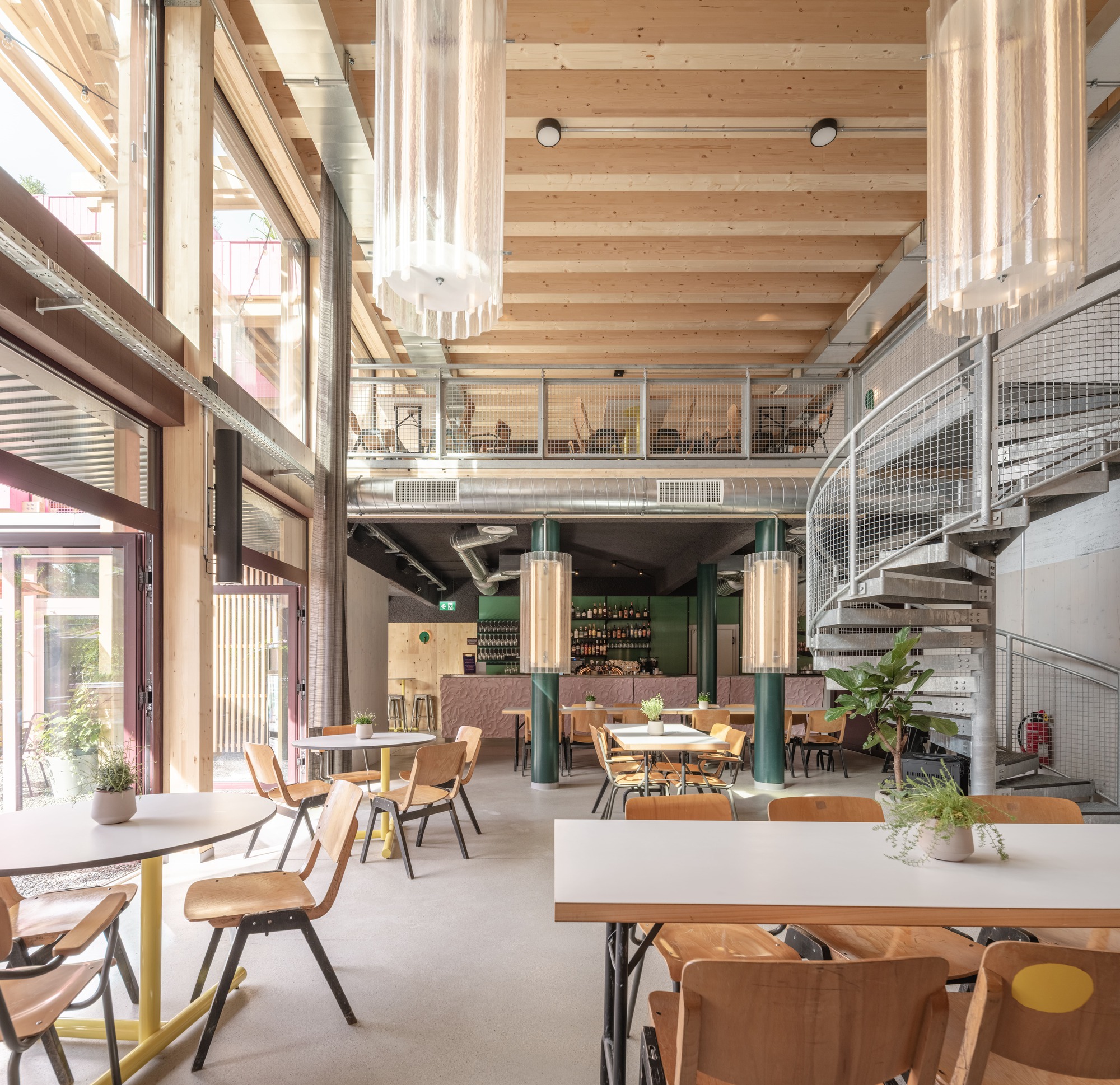 Double-height cafe interior with timber beam ceiling, green columns, spiral mesh stair and wooden chairs, daylight streaming through tall windows