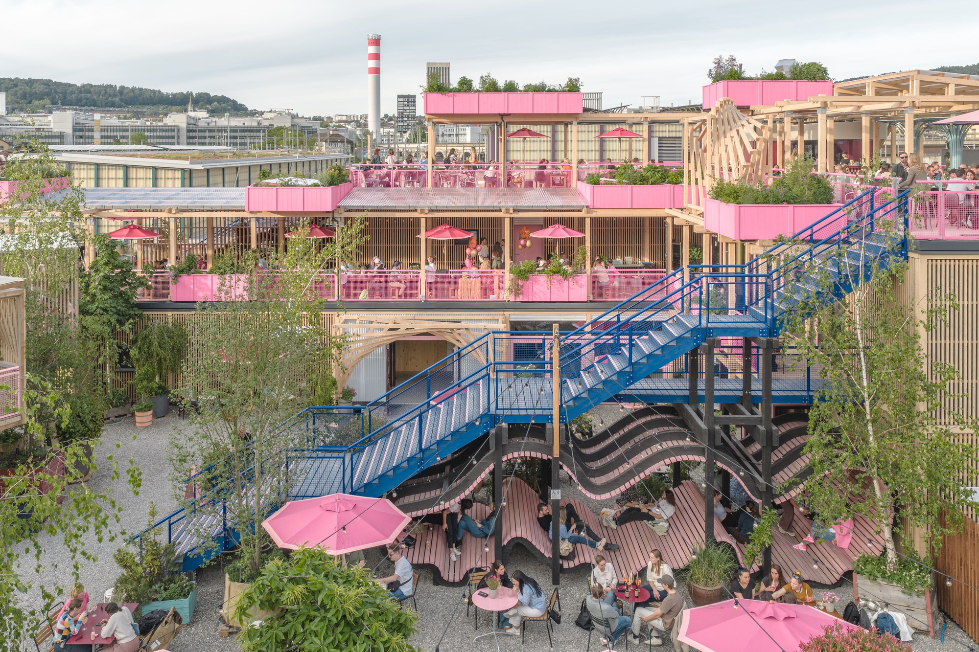 Elevated view of a blue steel staircase crossing pink-planted terraces above a gravel courtyard with umbrellas and seated diners at dusk