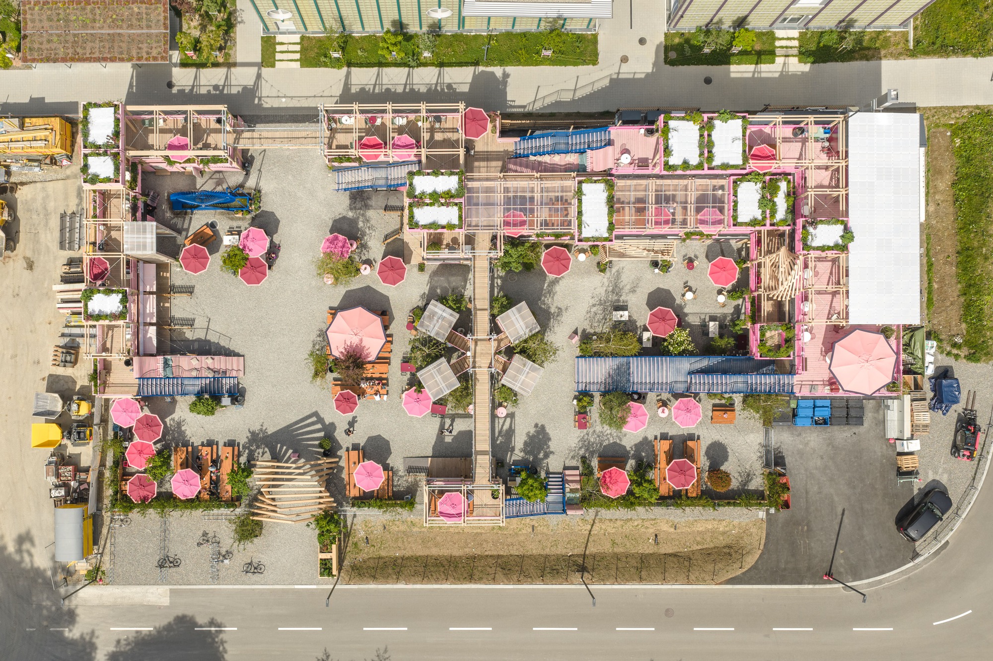 Top-down drone view of a timber pavilion and gravel courtyard dotted with pink umbrellas beside a street in bright daylight