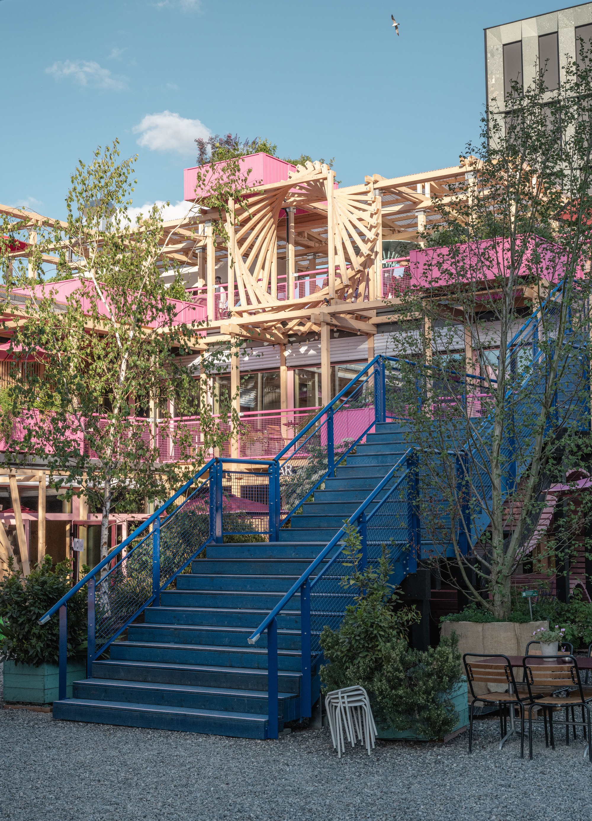 Blue steel staircase with mesh railings rising toward a timber fan-shaped frame and pink planters under a clear sky