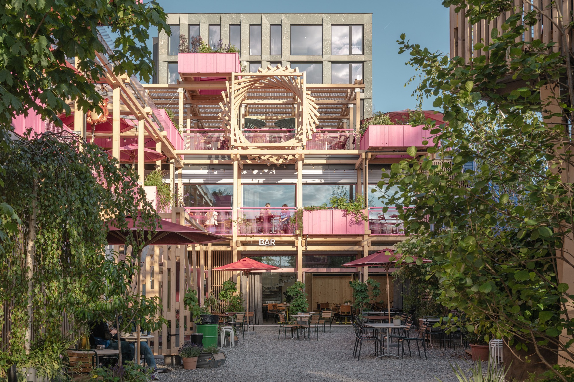 Courtyard facade with a circular timber fan detail above the bar sign, framed by birch leaves in warm evening light
