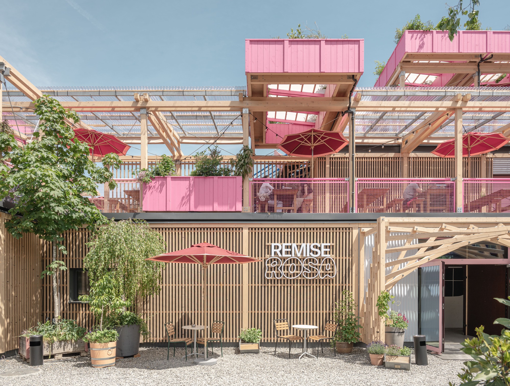 Slatted timber wall with the neon-style Remise Rosa sign beneath pink planters and red umbrellas over a gravel terrace