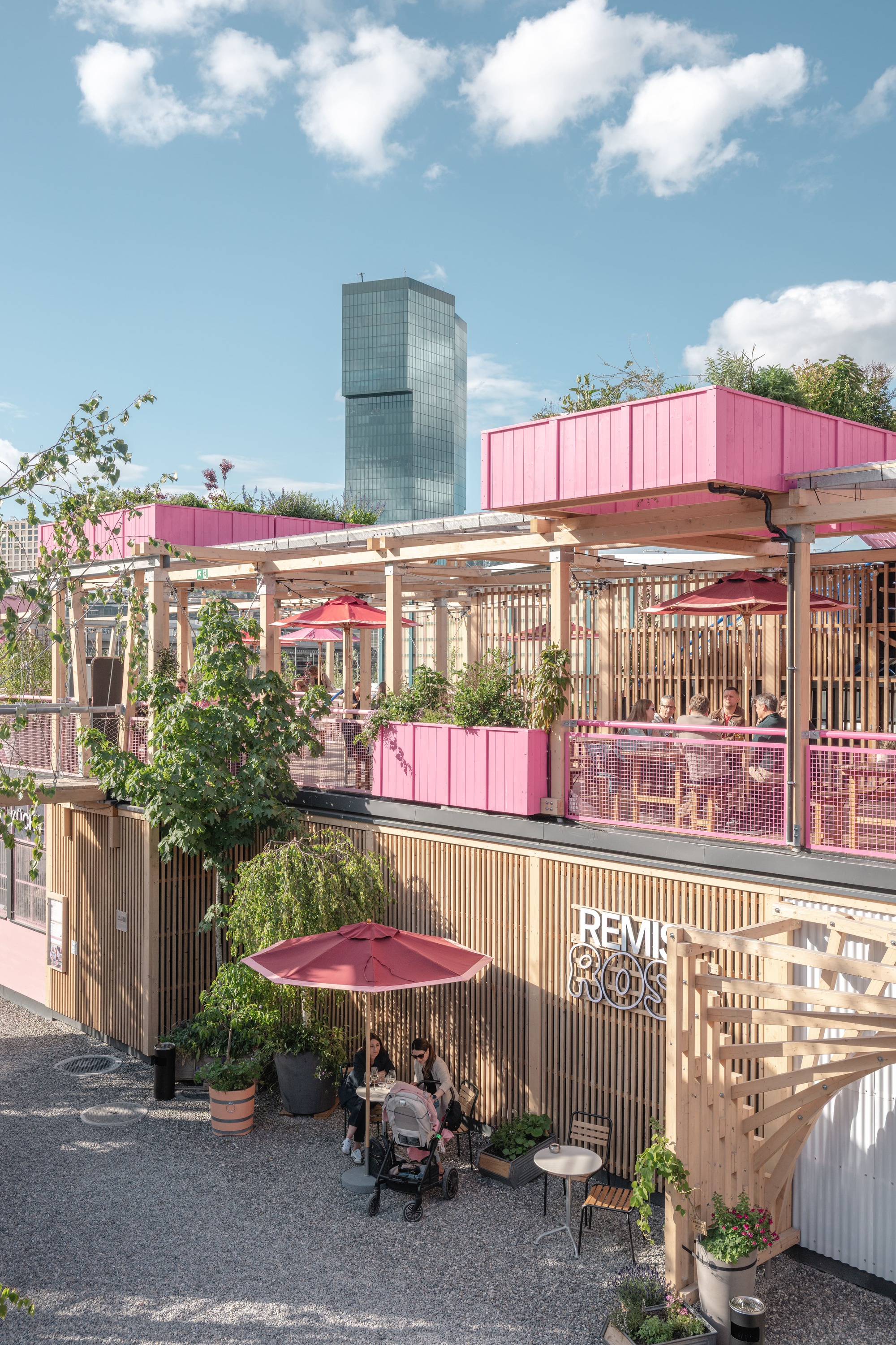 Timber-slatted pavilion with pink planter boxes and a rooftop terrace, red parasols and diners below a glass tower in afternoon sun