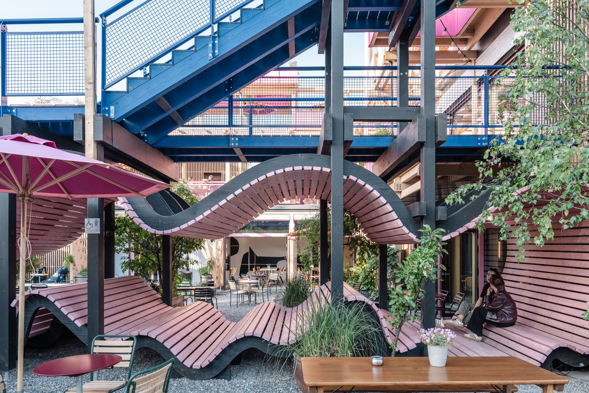 Undulating pink slatted bench winding between dark timber columns beneath a blue steel staircase, with a couple seated on the right