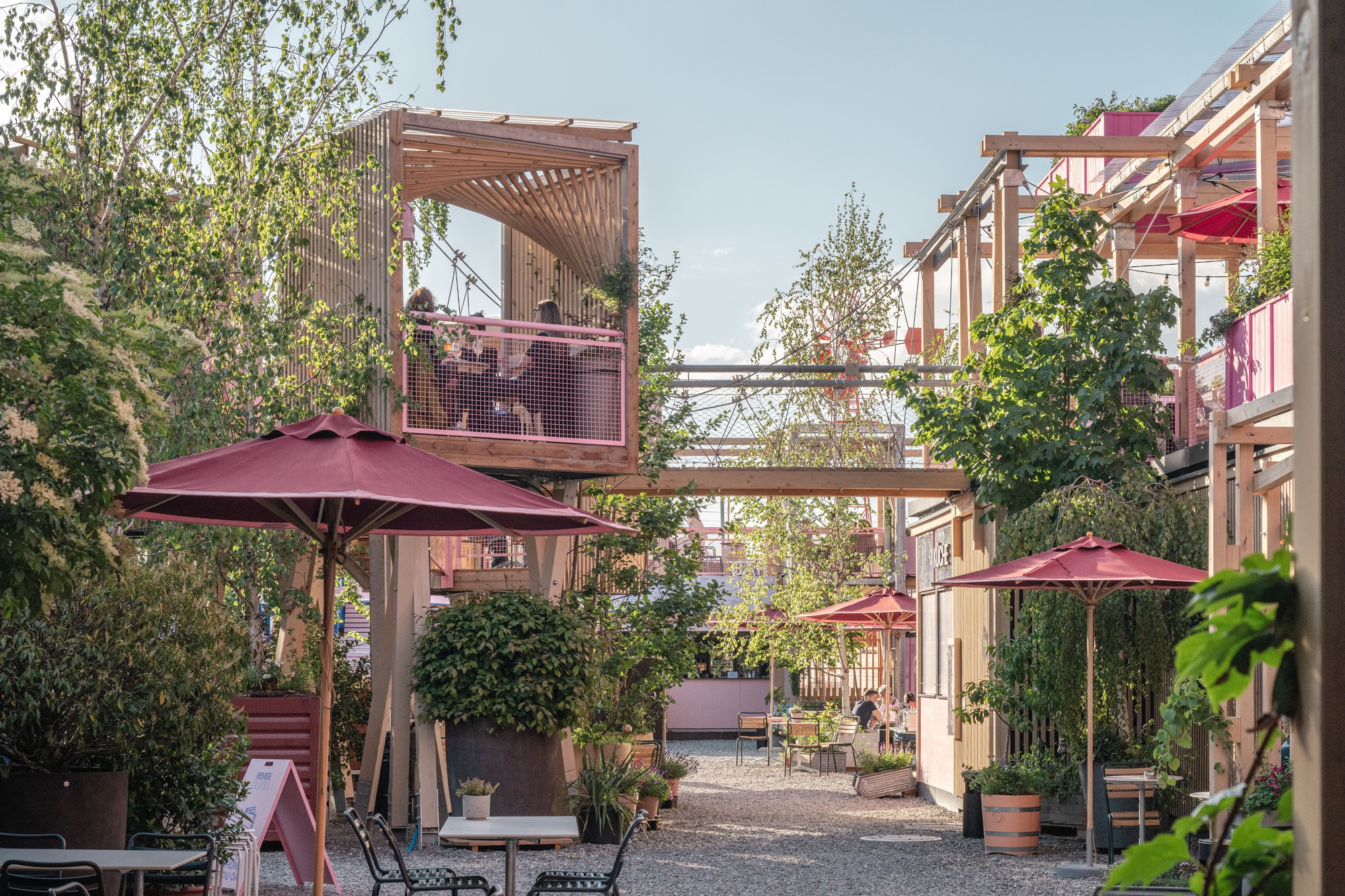 Gravel lane lined with red parasols, potted trees and timber balconies, where people sit on an elevated deck in warm evening light