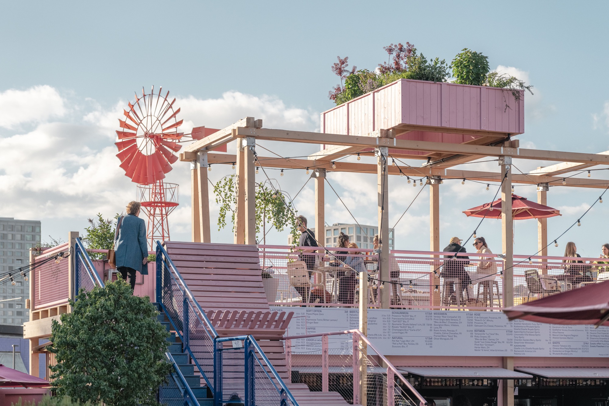 Pink stair with blue railings rising to a rooftop terrace with a red windmill, string lights and diners under scattered clouds