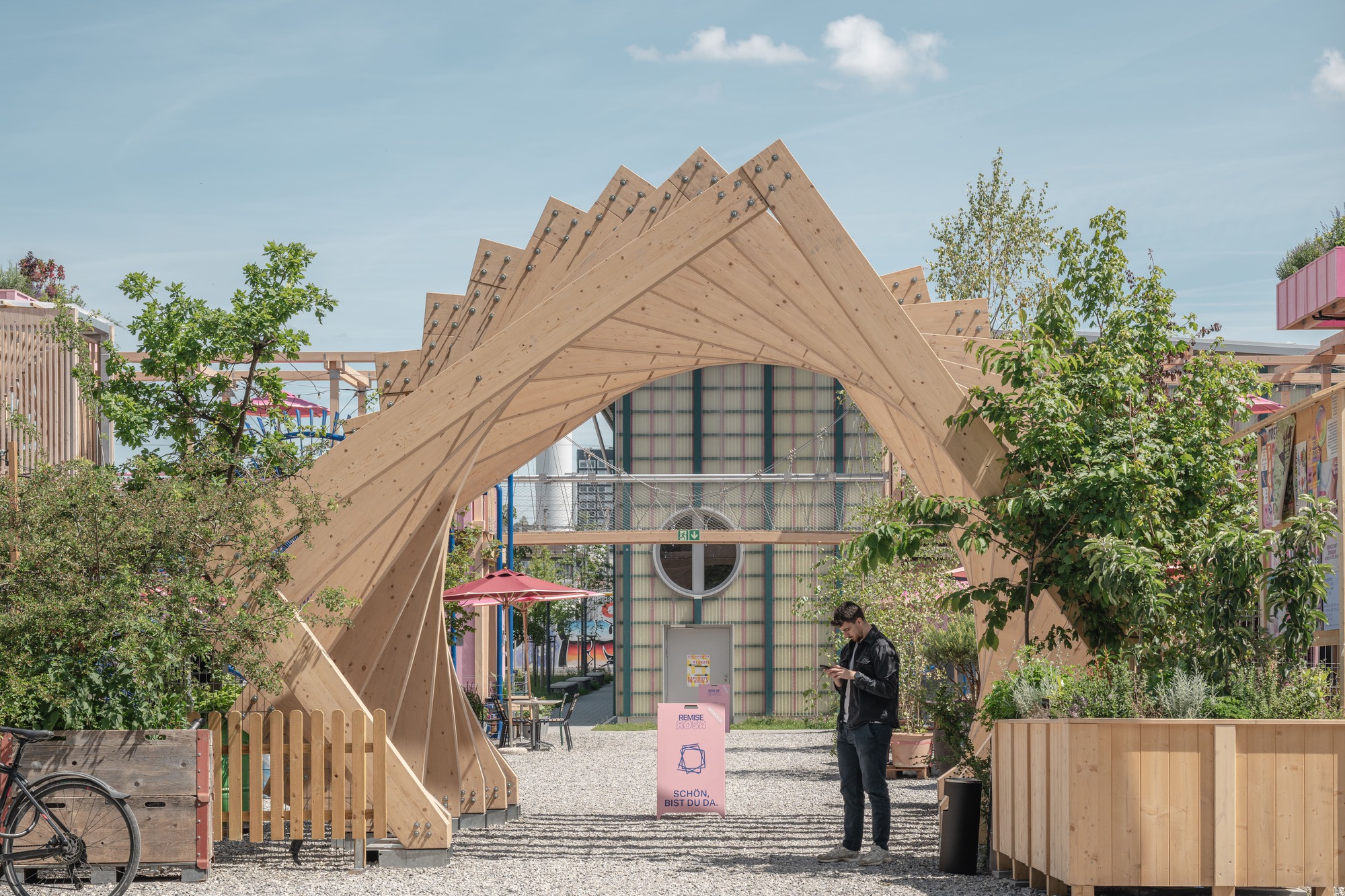 Layered timber arch framing a glazed hall beyond, with a man checking his phone on the gravel beside planter boxes