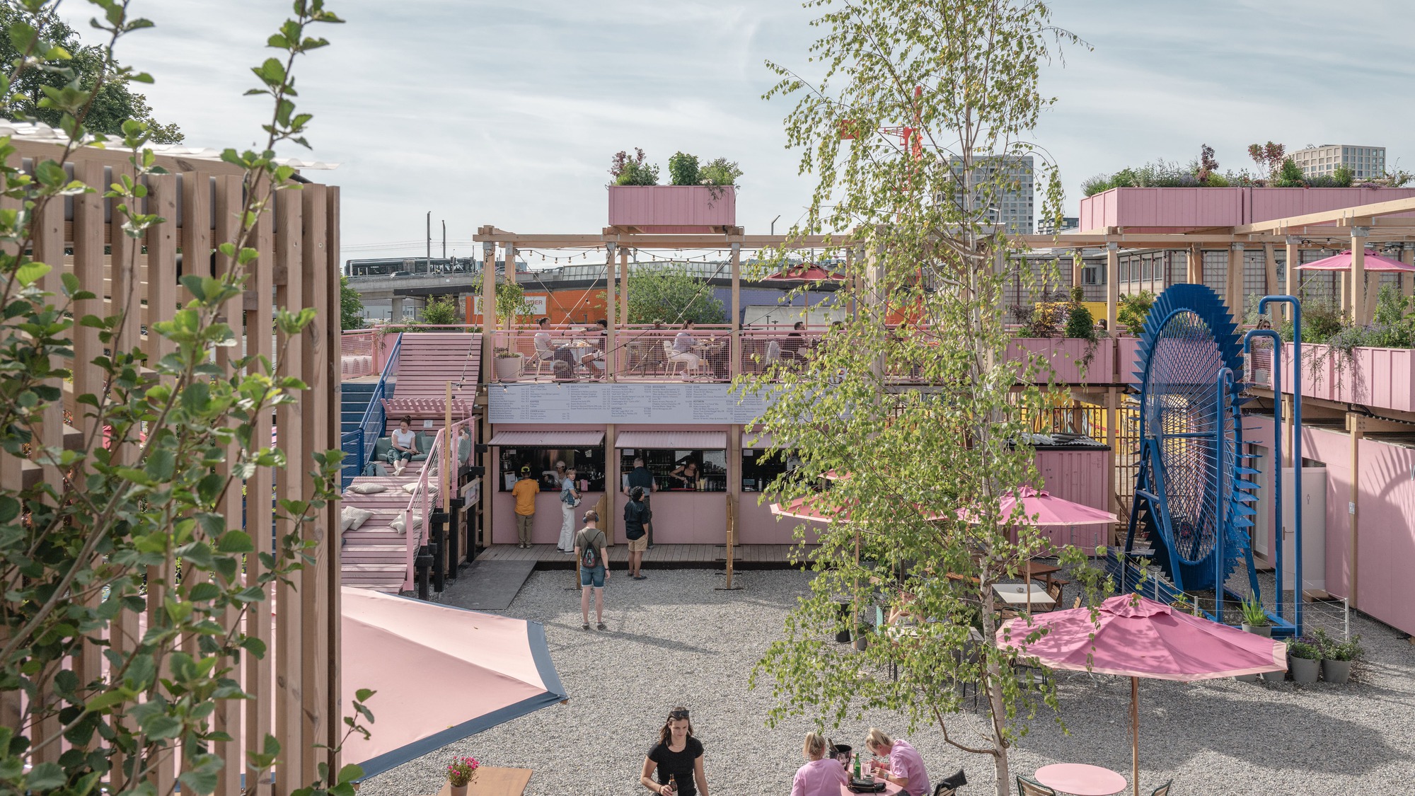 Gravel courtyard with pink umbrellas, a blue wheel structure and timber terraces, seen through birch leaves on a bright day