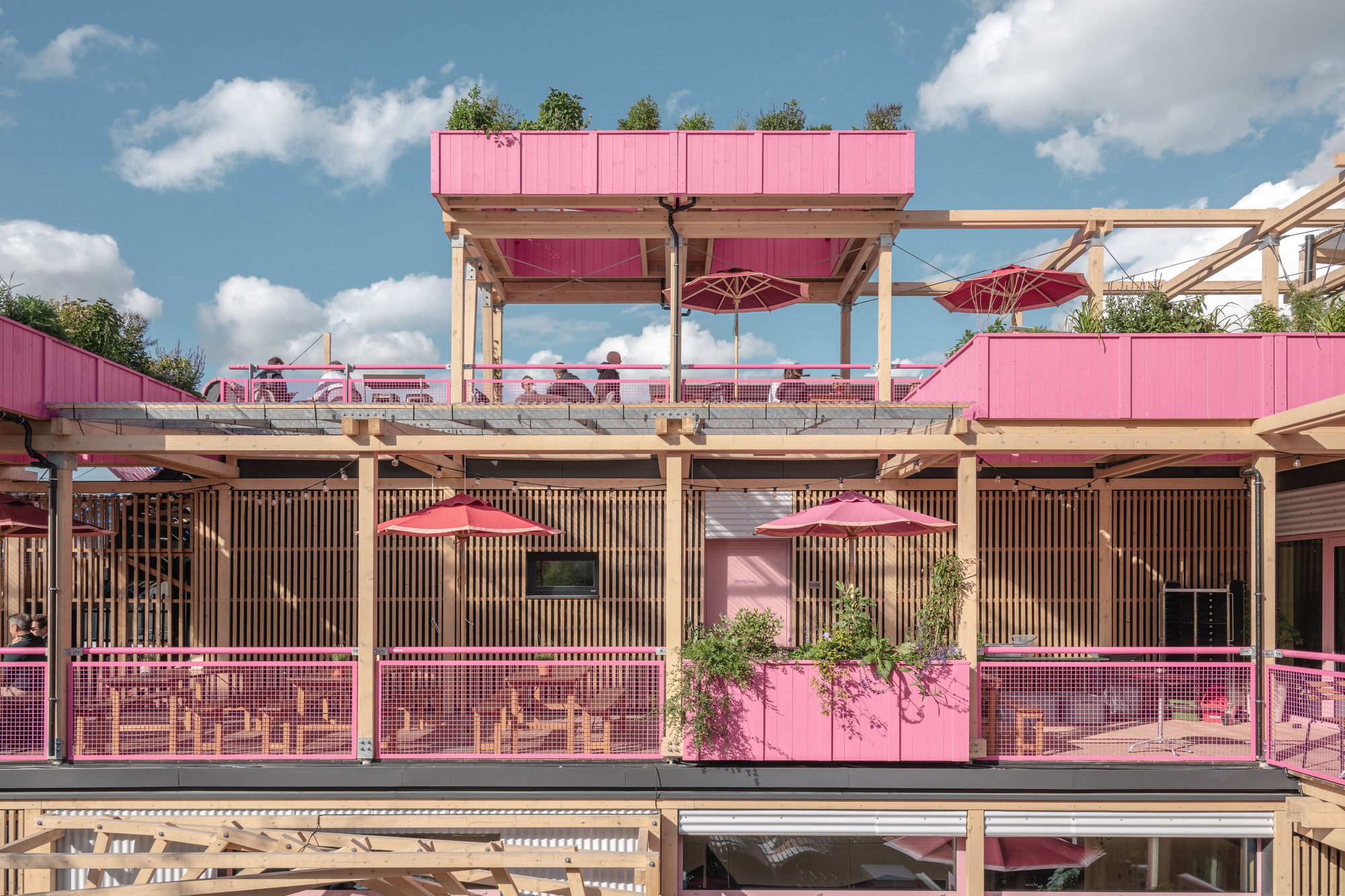 Pink planter-topped terraces with timber frame, red parasols and railings, with diners at tables under scattered clouds