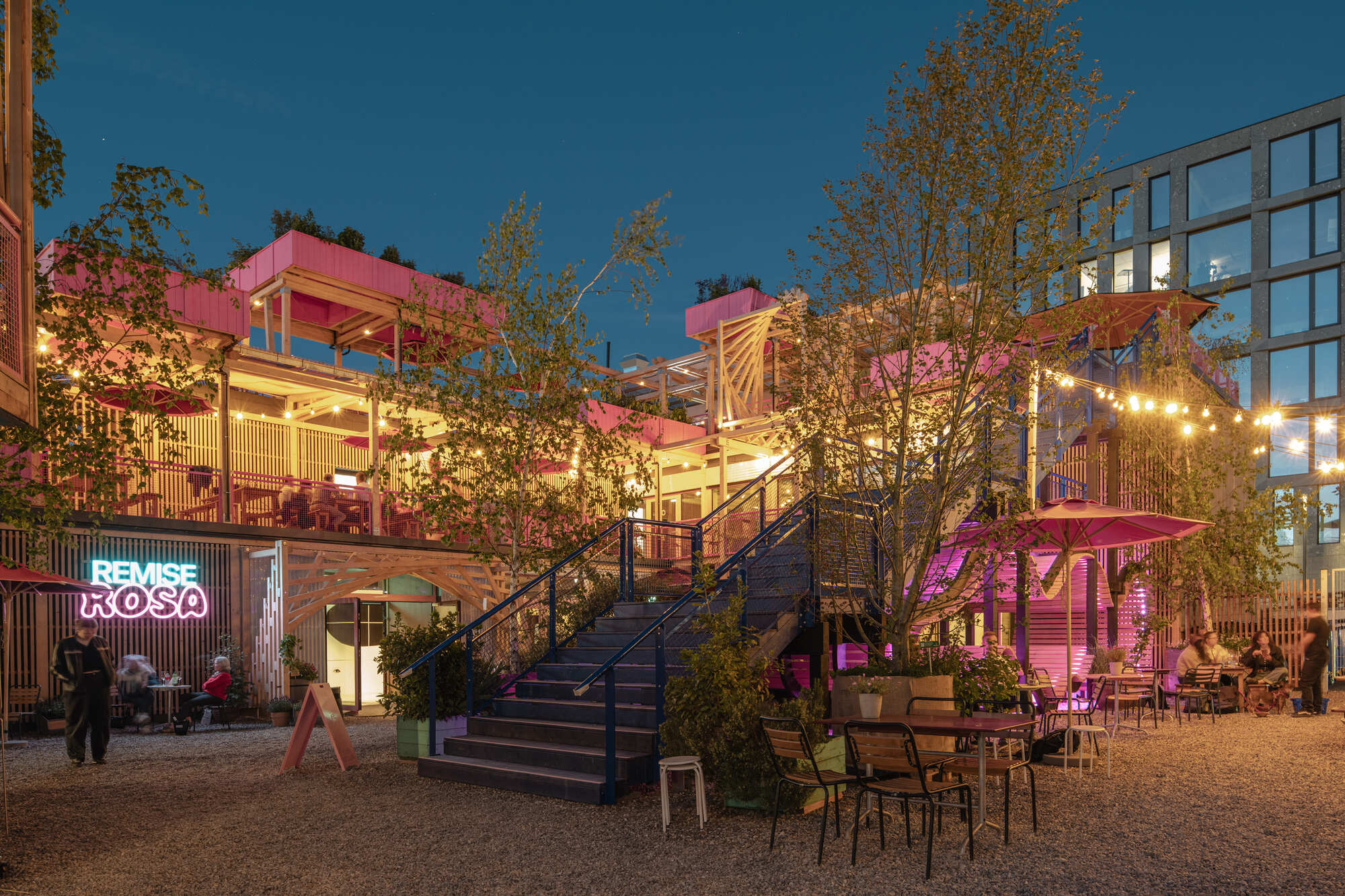 Dusk view of a blue steel staircase rising through the timber and pink complex, with string lights and a neon sign