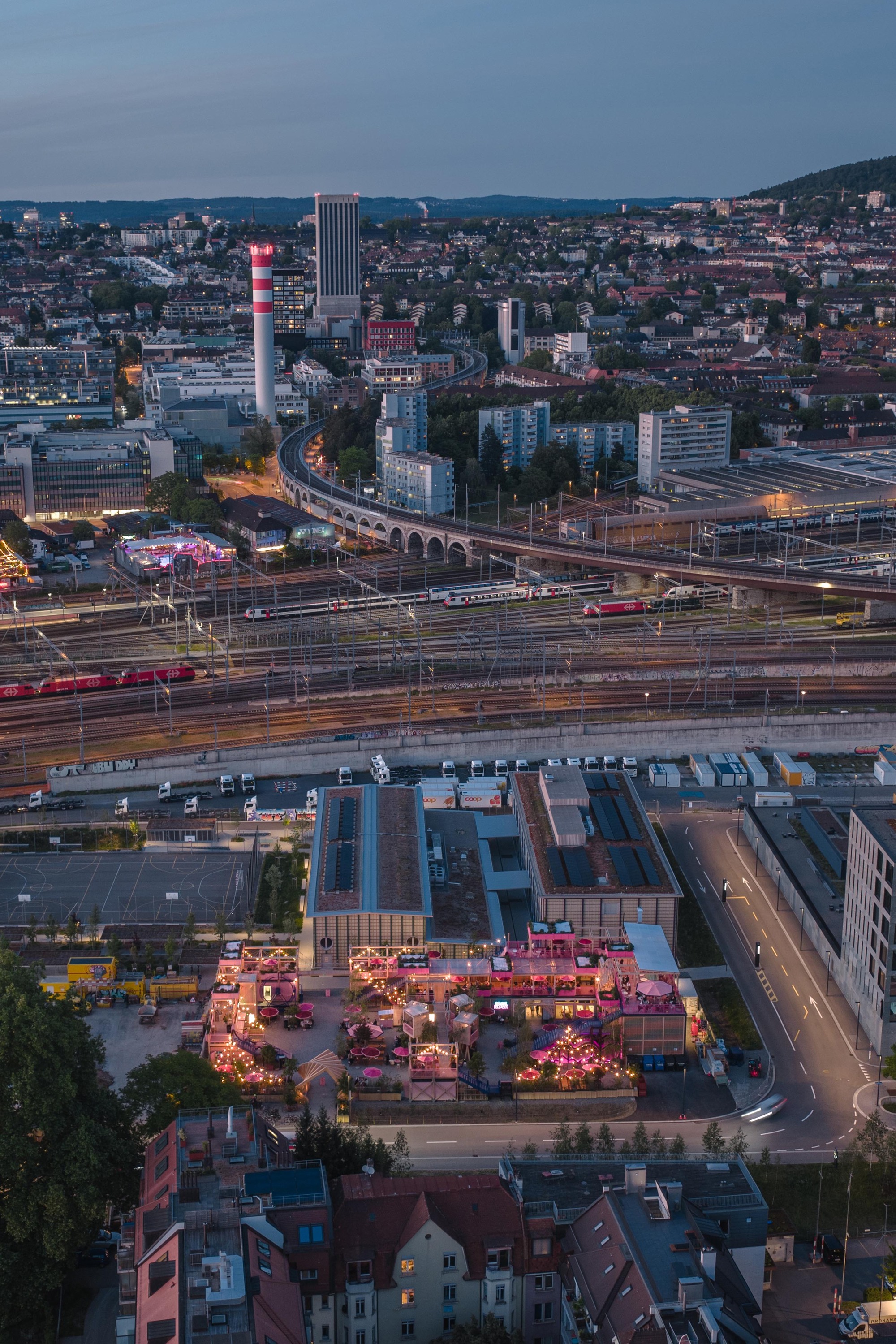 Aerial view at dusk of the lit pink terraced complex beside rail tracks, with a striped chimney and viaduct beyond