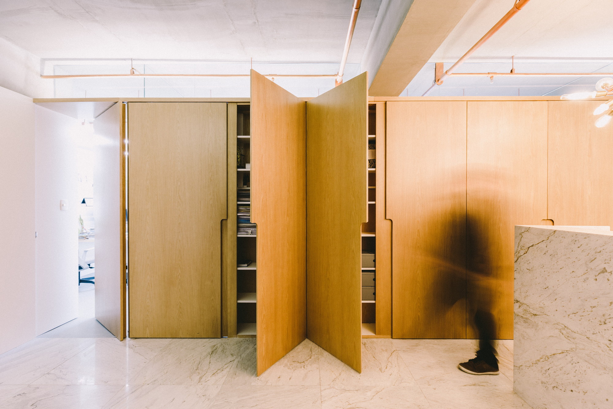 Oak storage wall with pivoting doors swung open onto shelves, a blurred person walking past a marble counter