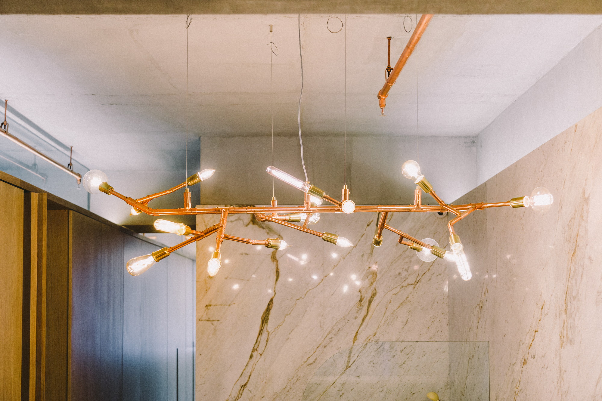 Branching copper pipe chandelier with bare filament bulbs hanging in front of a veined marble slab wall