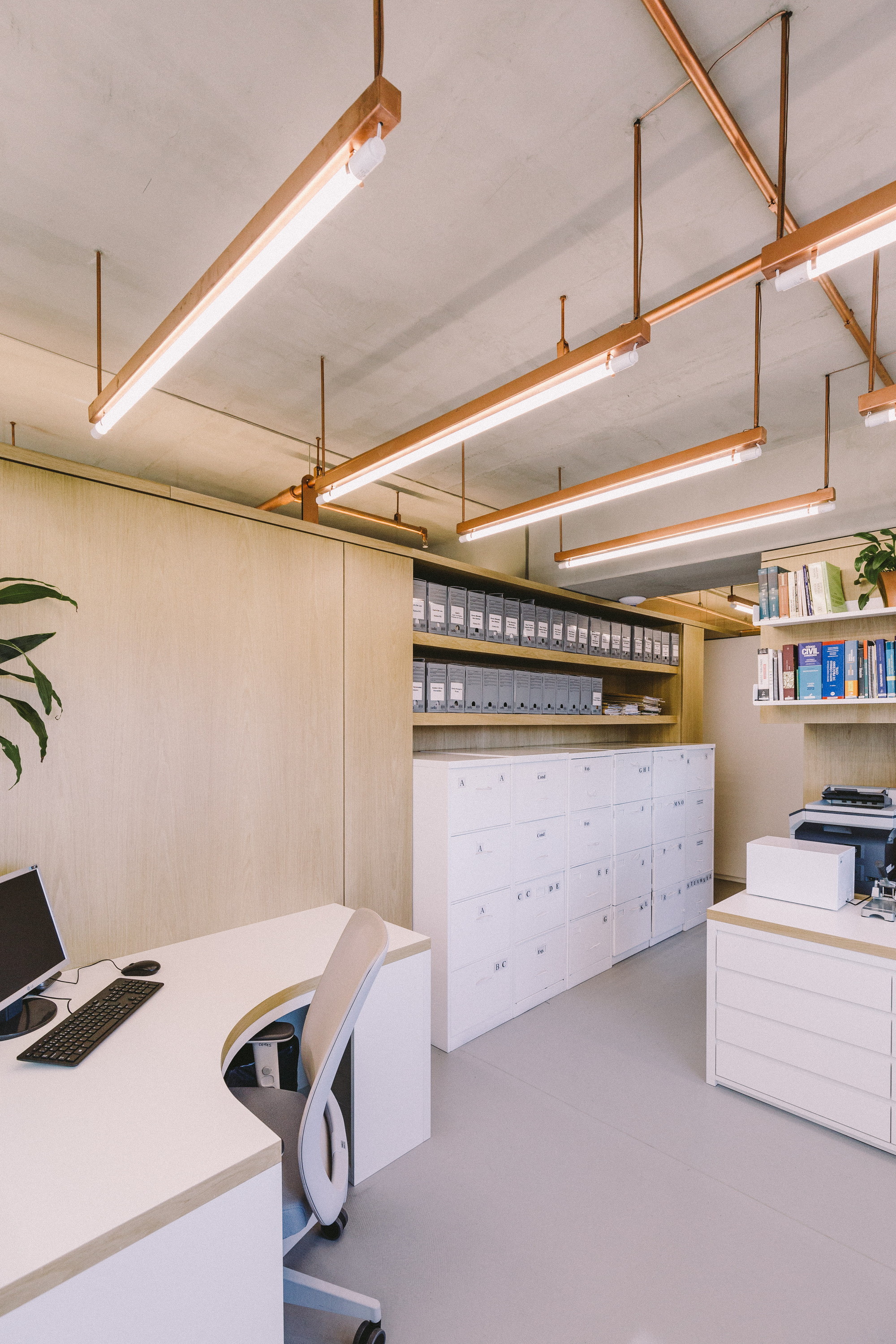 Office corner with curved desk, filing cabinets and archive boxes on oak shelving beneath suspended copper light fixtures in daylight
