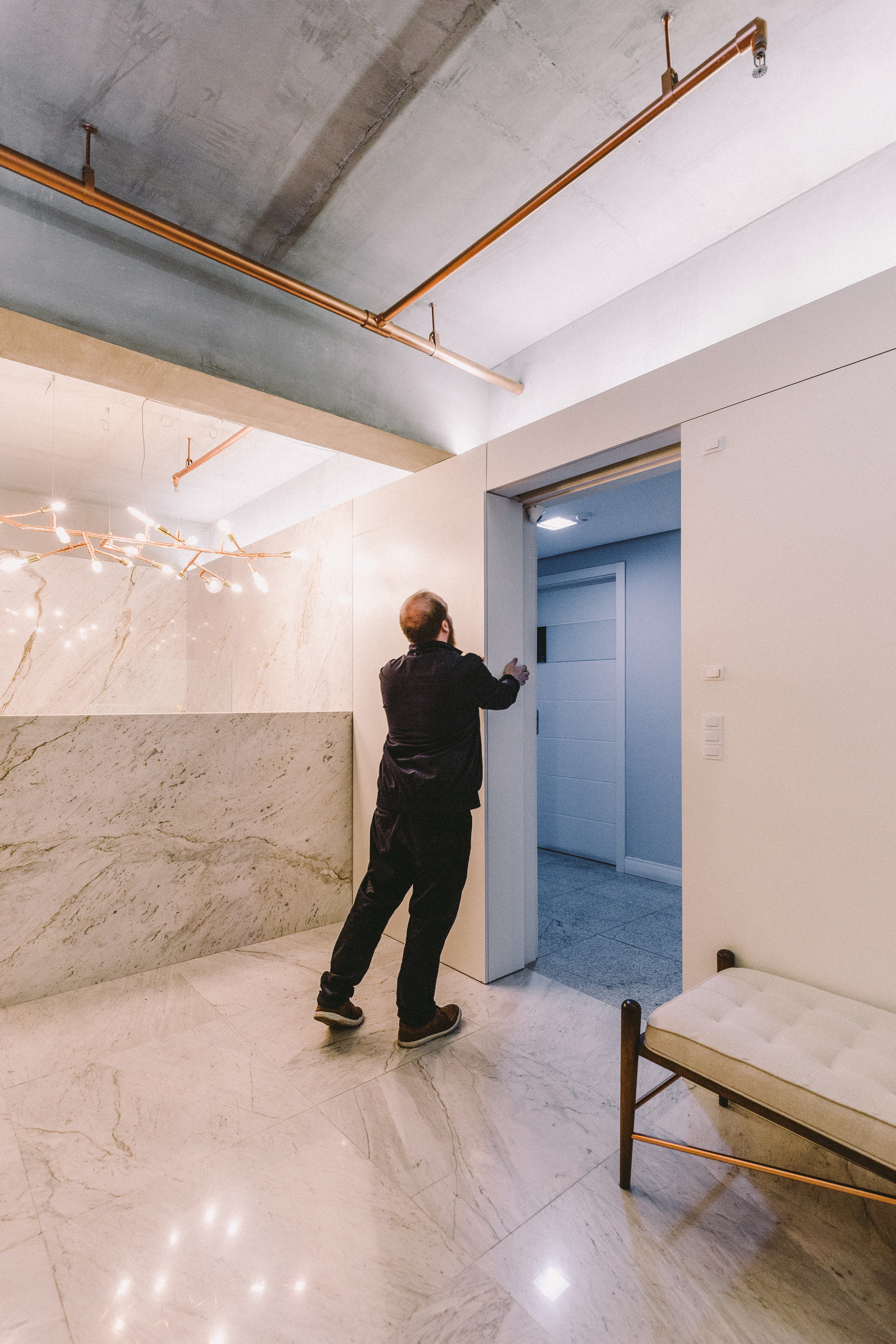 Man pushing open a tall white pivot door in a marble-clad entry hall under copper conduits and a branching light fixture