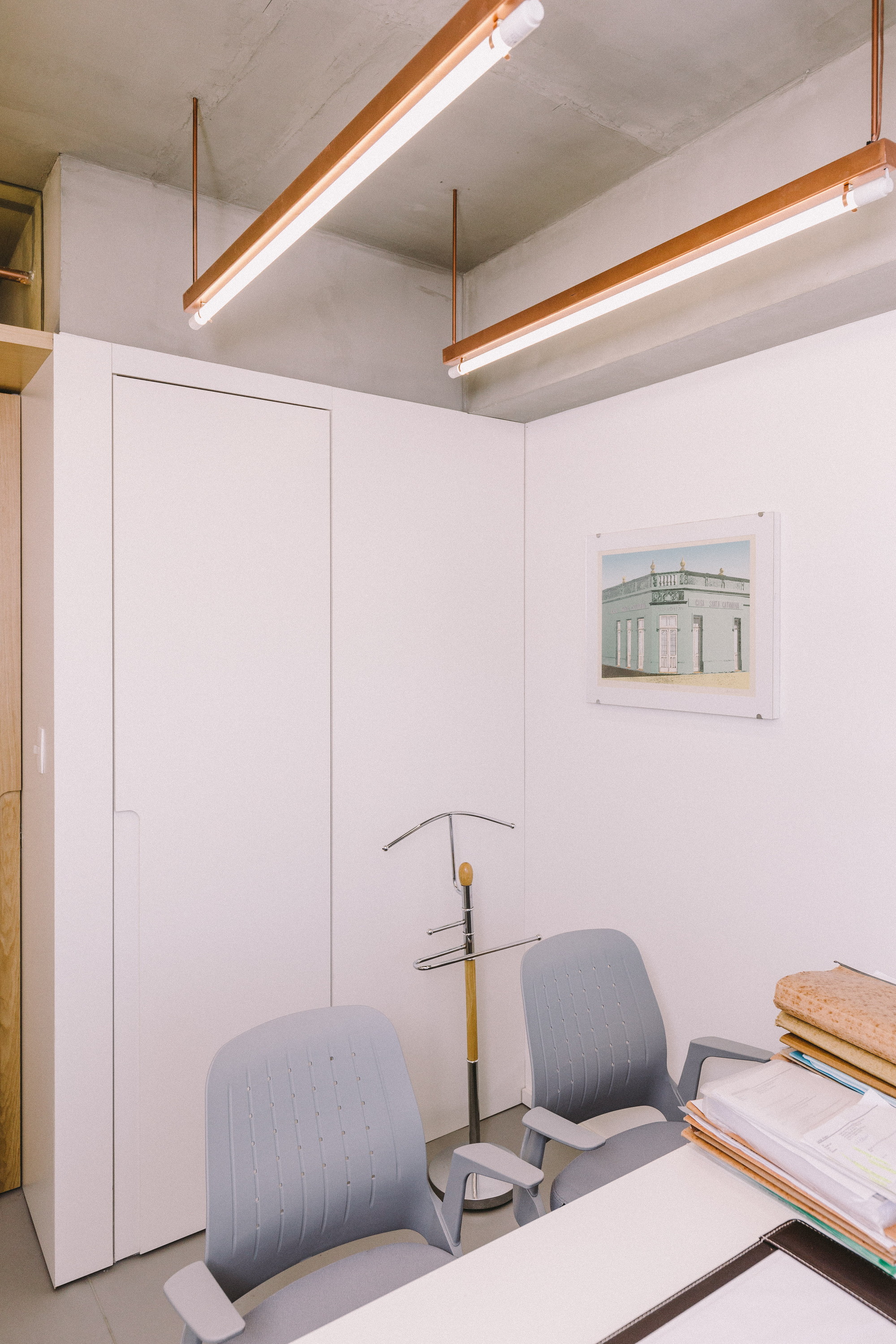 Corner of an office with white built-in cabinets, grey chairs and a framed photograph beneath copper-trimmed tube lights