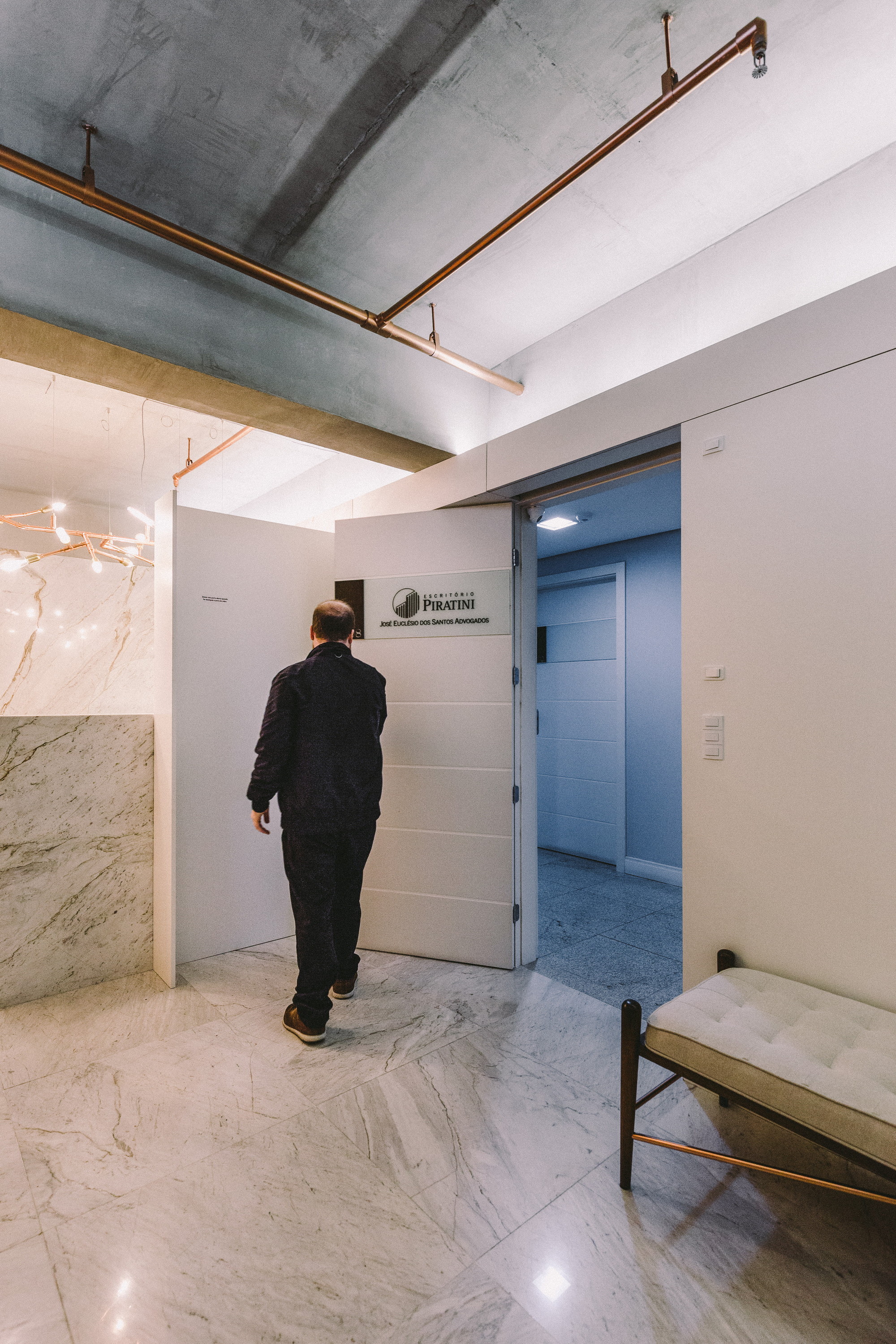 Visitor walking through the opened white entry door with a firm nameplate, past a marble wall and tufted bench