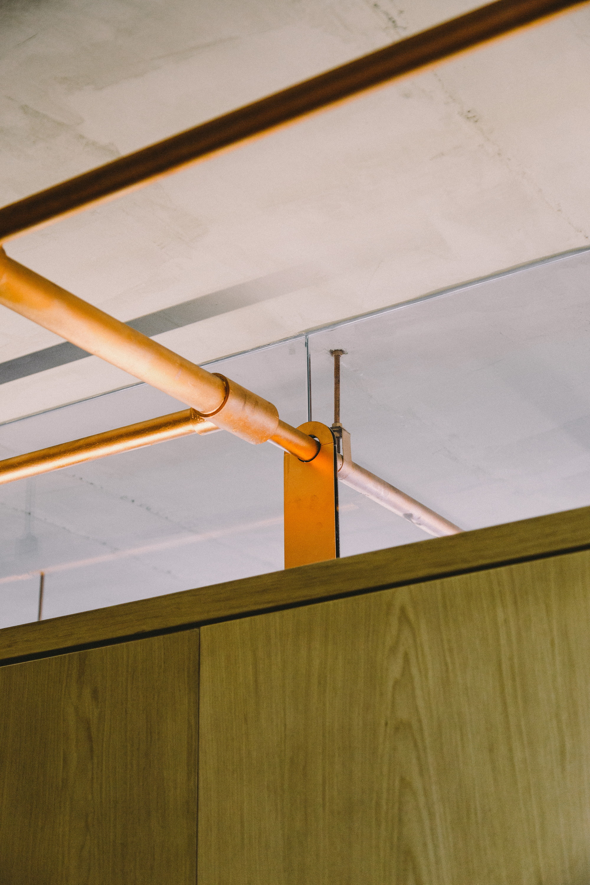 Close-up of copper pipes on an orange steel bracket above an oak-veneer cabinet and exposed concrete ceiling